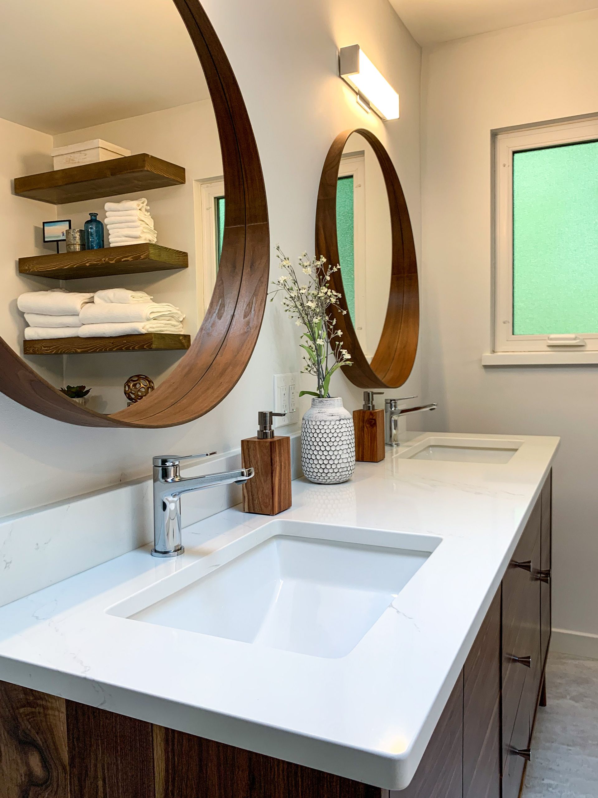 Modern bathroom with double vanity, two round wooden mirrors, and open shelves with folded towels.