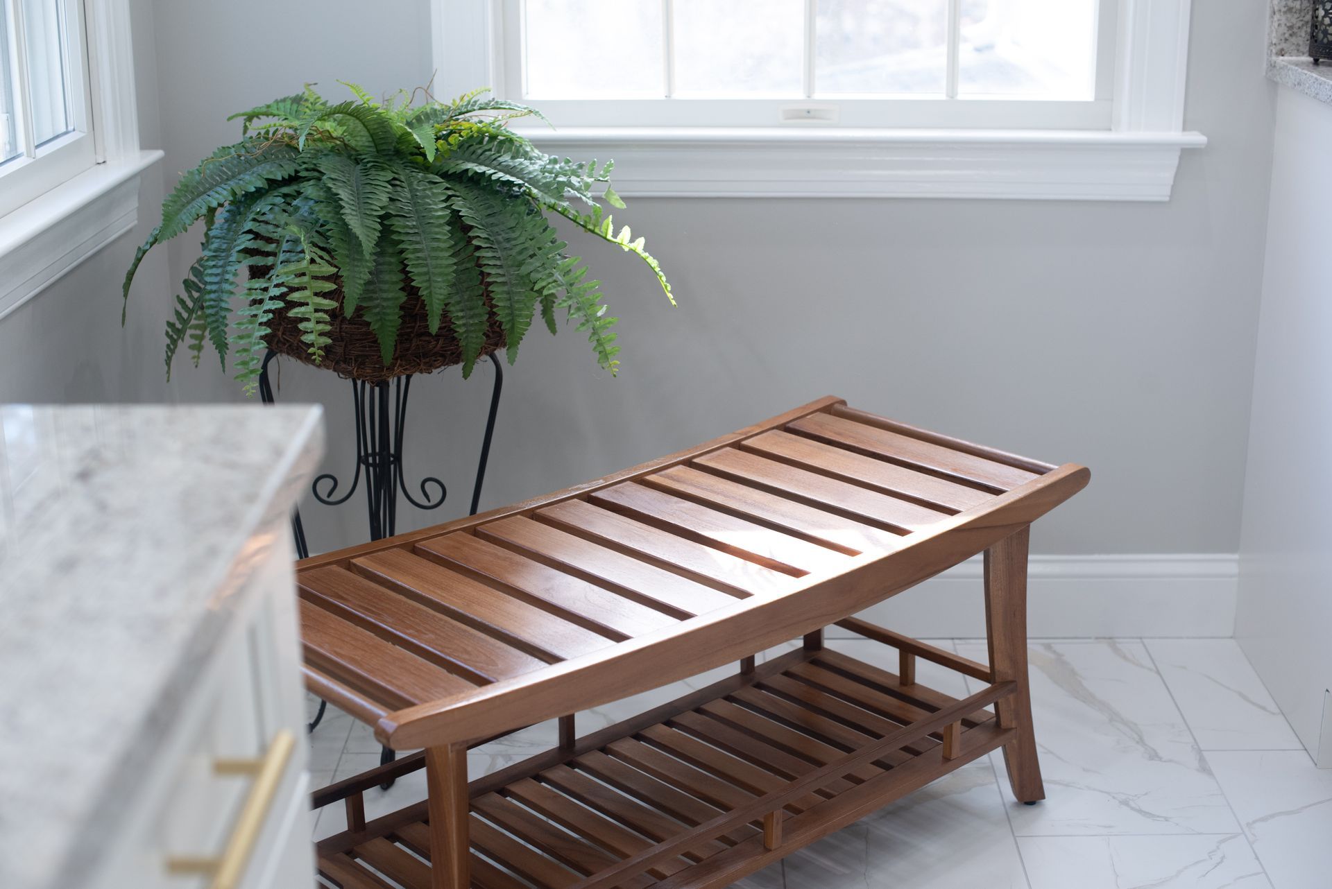 A wooden bench sits in a bathroom with a fern plant by the window. The walls are gray, and the floor is tiled.