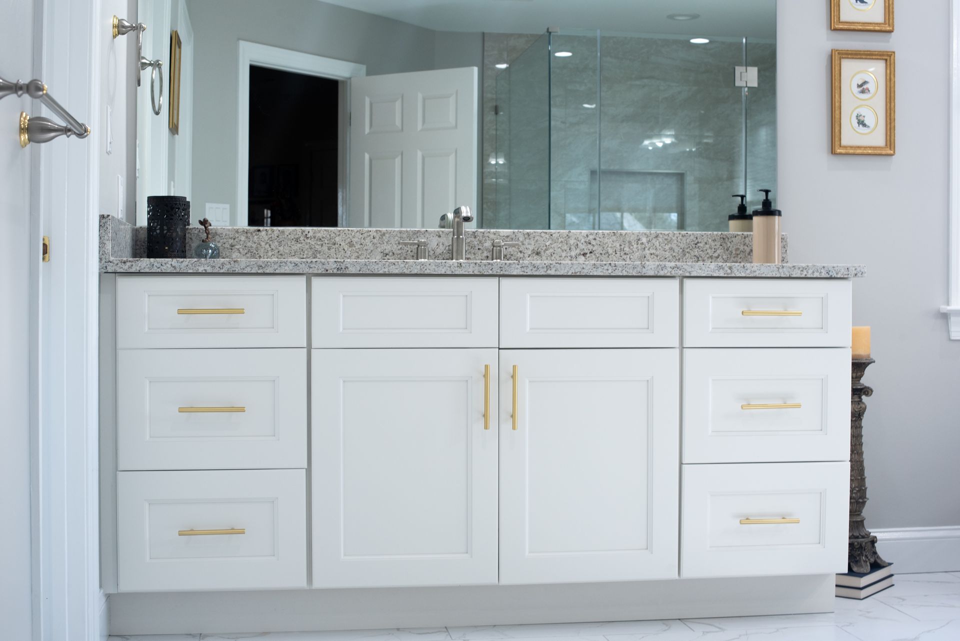 A white bathroom vanity with gold hardware and a speckled countertop. A large mirror reflects a shower and door.