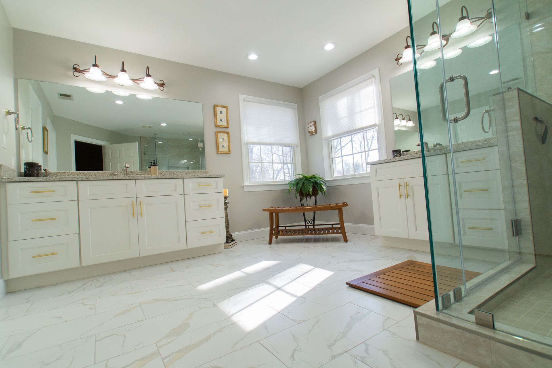 Spacious, light-filled bathroom with white cabinetry, large mirror, and glass shower. Features include marble-look flooring, windows, and a wooden bench.