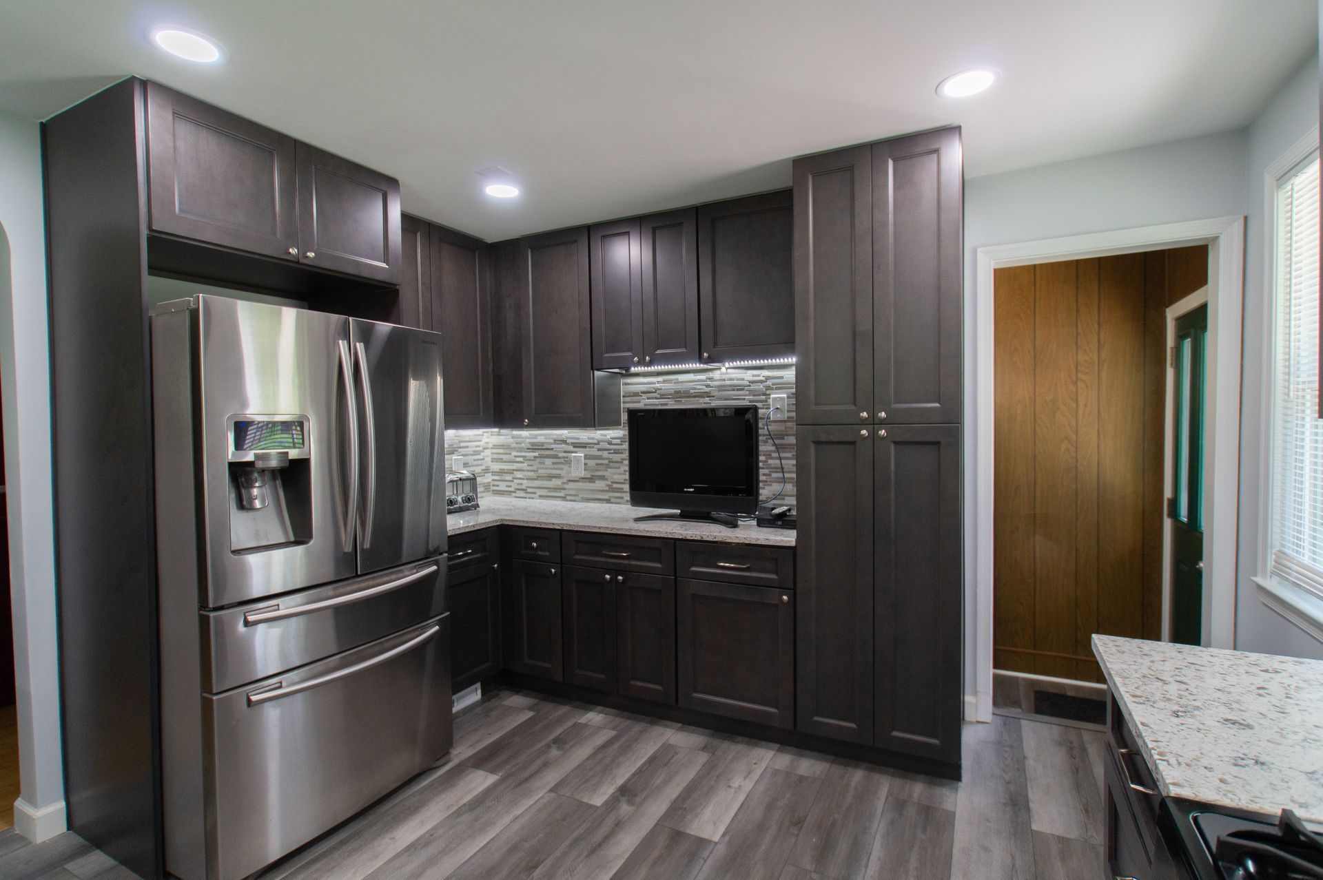 Modern kitchen with dark brown cabinets, stainless steel refrigerator, and light gray wood-look flooring. A television is mounted on the backsplash.