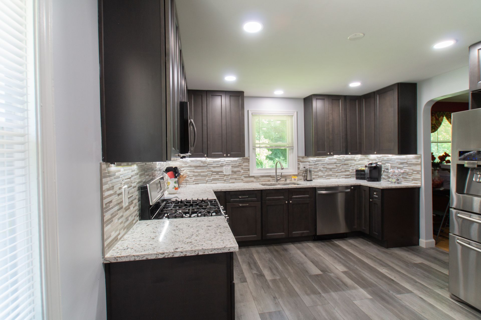 Modern kitchen with dark brown cabinets, granite countertops, stainless steel appliances, and gray wood-look flooring.