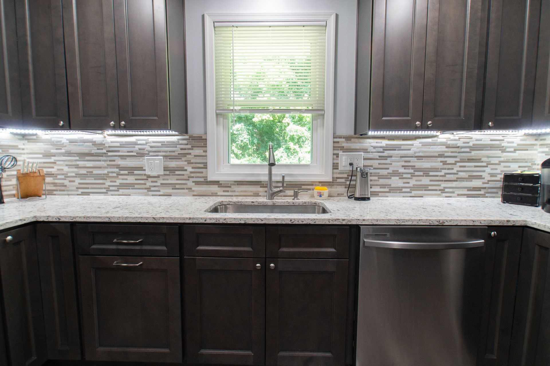 Kitchen with dark brown cabinets, light-colored speckled countertops, and a textured backsplash. A window with blinds is above the sink.