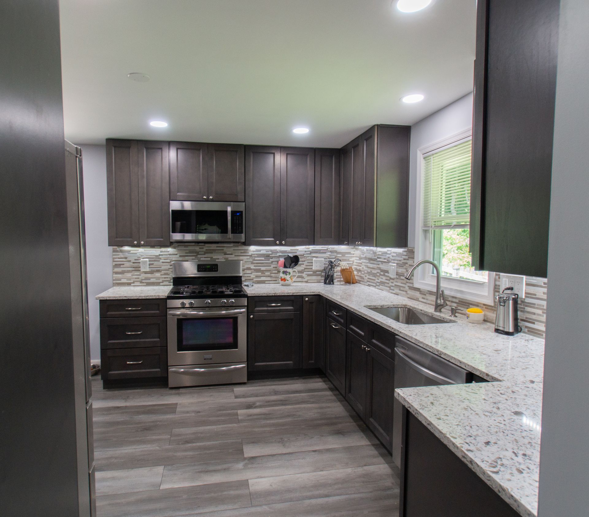 Dark-toned kitchen with wood-look flooring, dark cabinets, stainless steel appliances, and a gray speckled countertop.