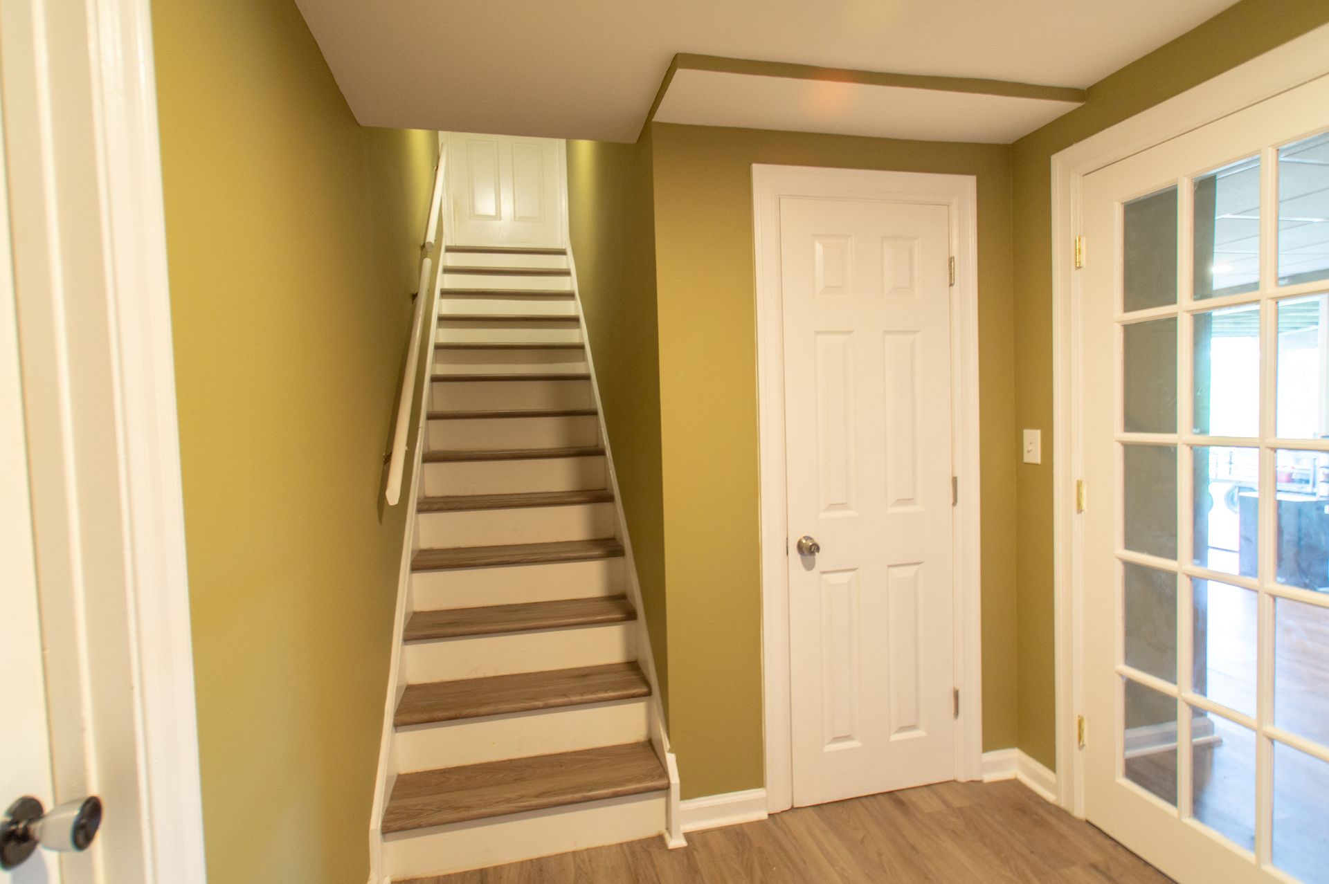 Staircase with light brown steps leading up to a white door and ceiling access. Light green walls and white trim surround.