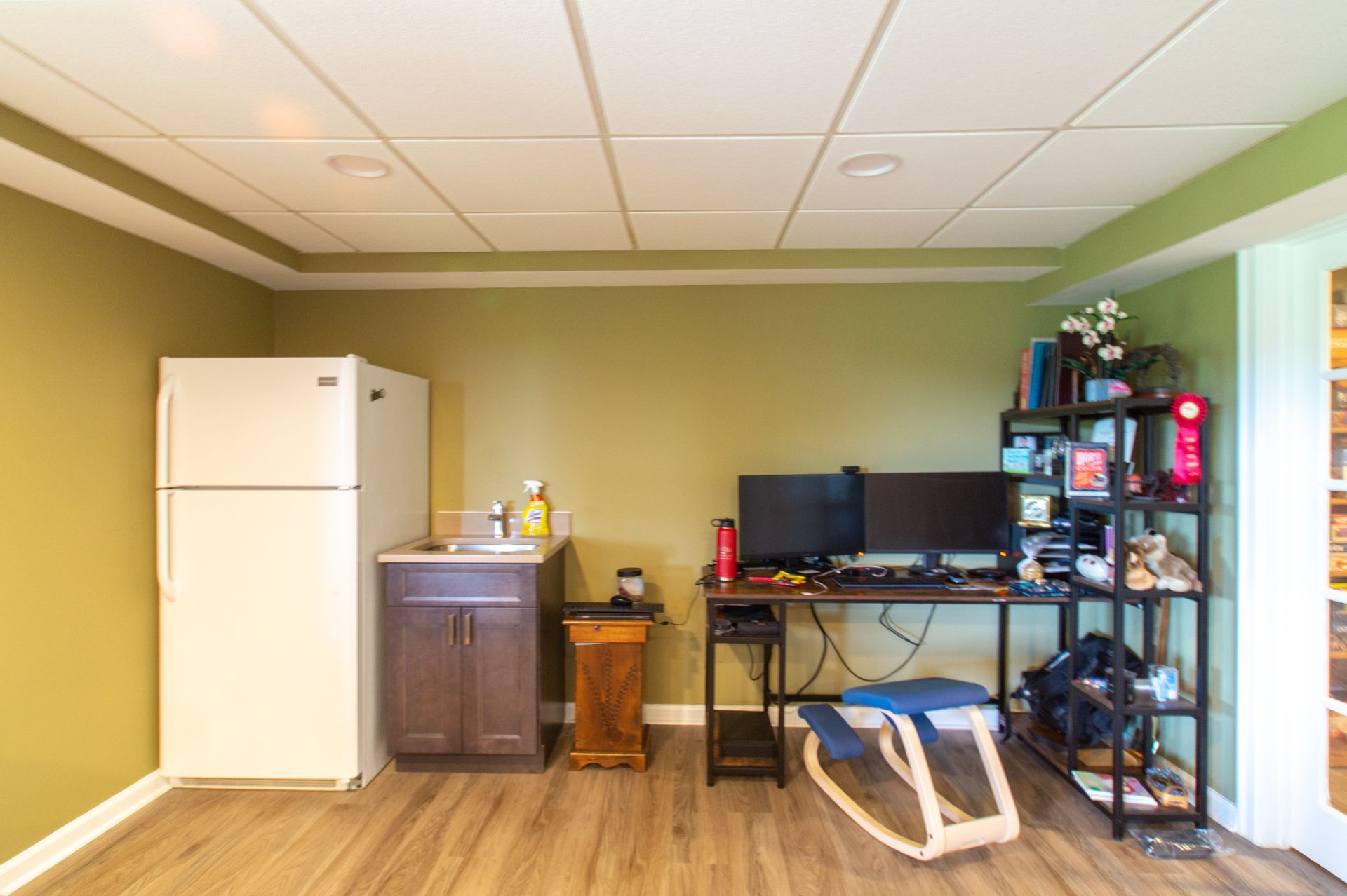 A home office with a refrigerator, desk with dual monitors, and a small sink. Green walls and wood-look flooring.
