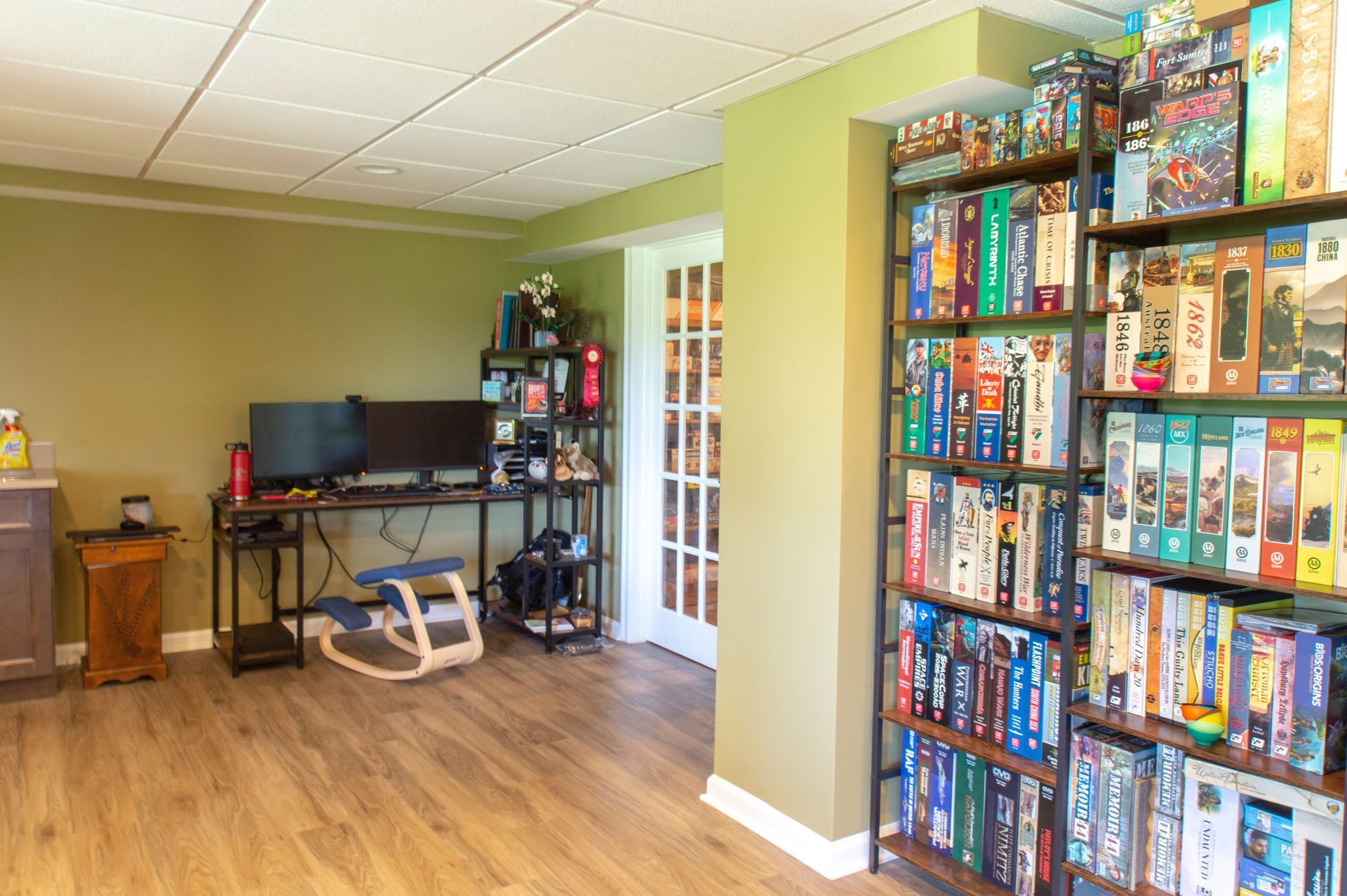 A game room with a desk, shelves full of board games, and double doors. Olive green walls and faux wood flooring.