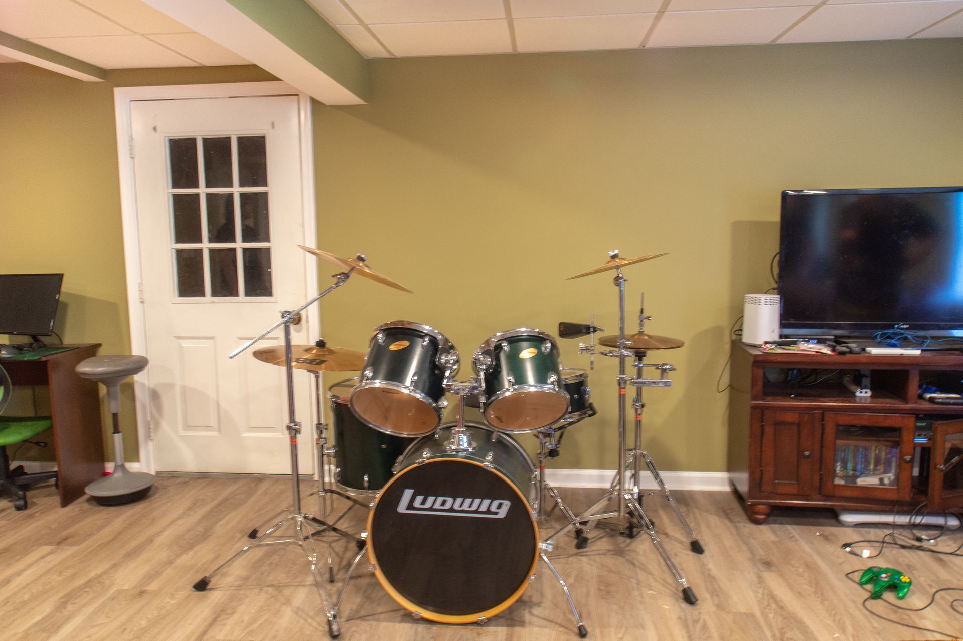 A drum set in a basement, facing a door and TV.  The set has black drums and cymbals, against a green wall and light wood floor.