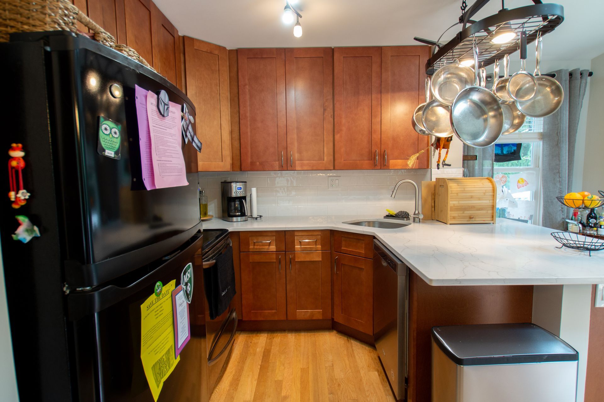A kitchen with brown cabinets, a black refrigerator, light countertops, and a rack of pots hanging above the sink.