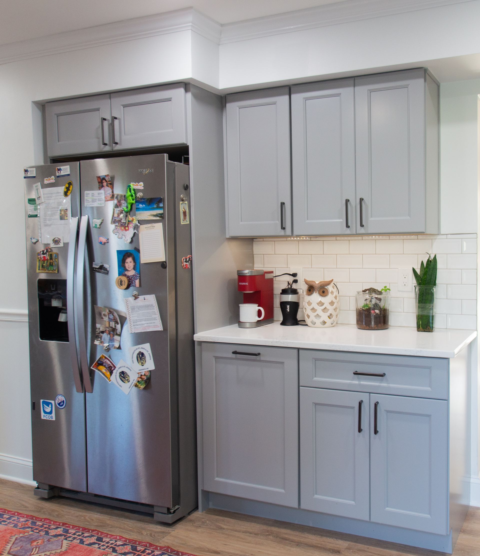 A kitchen corner with a stainless steel refrigerator, gray cabinets, and a white countertop. The corner has a coffee maker and some decor.