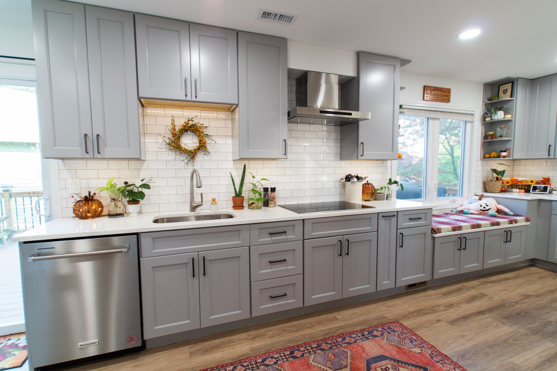 Gray kitchen with upper and lower cabinets, white countertops, and a stainless steel dishwasher. Includes a white tile backsplash, a wreath, and a window looking out onto a deck.