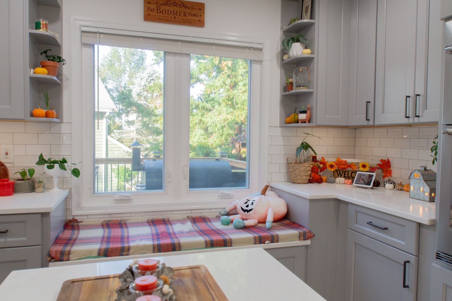 Cozy kitchen with grey cabinets, window seat, and fall decorations. A stuffed animal sits on the window seat with a plaid blanket.