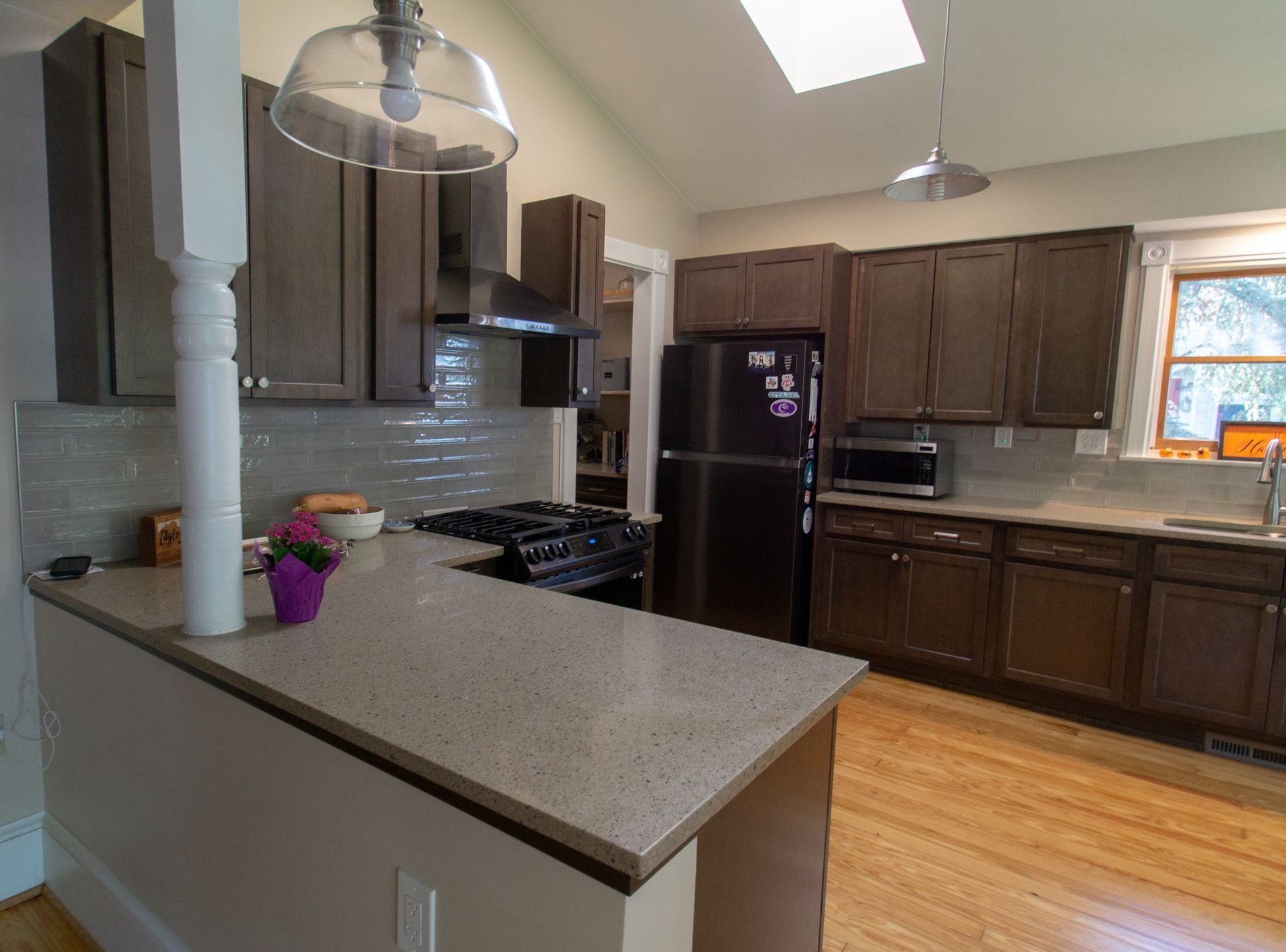 A kitchen with brown cabinets, a black refrigerator, and a light-colored countertop. Natural light streams in from a window and skylight.
