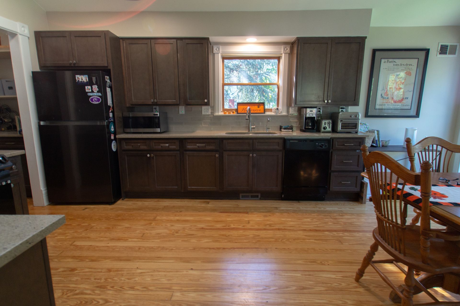 Kitchen with dark cabinets, stainless steel refrigerator, and light wood floors. A dining table and artwork are visible on the right.