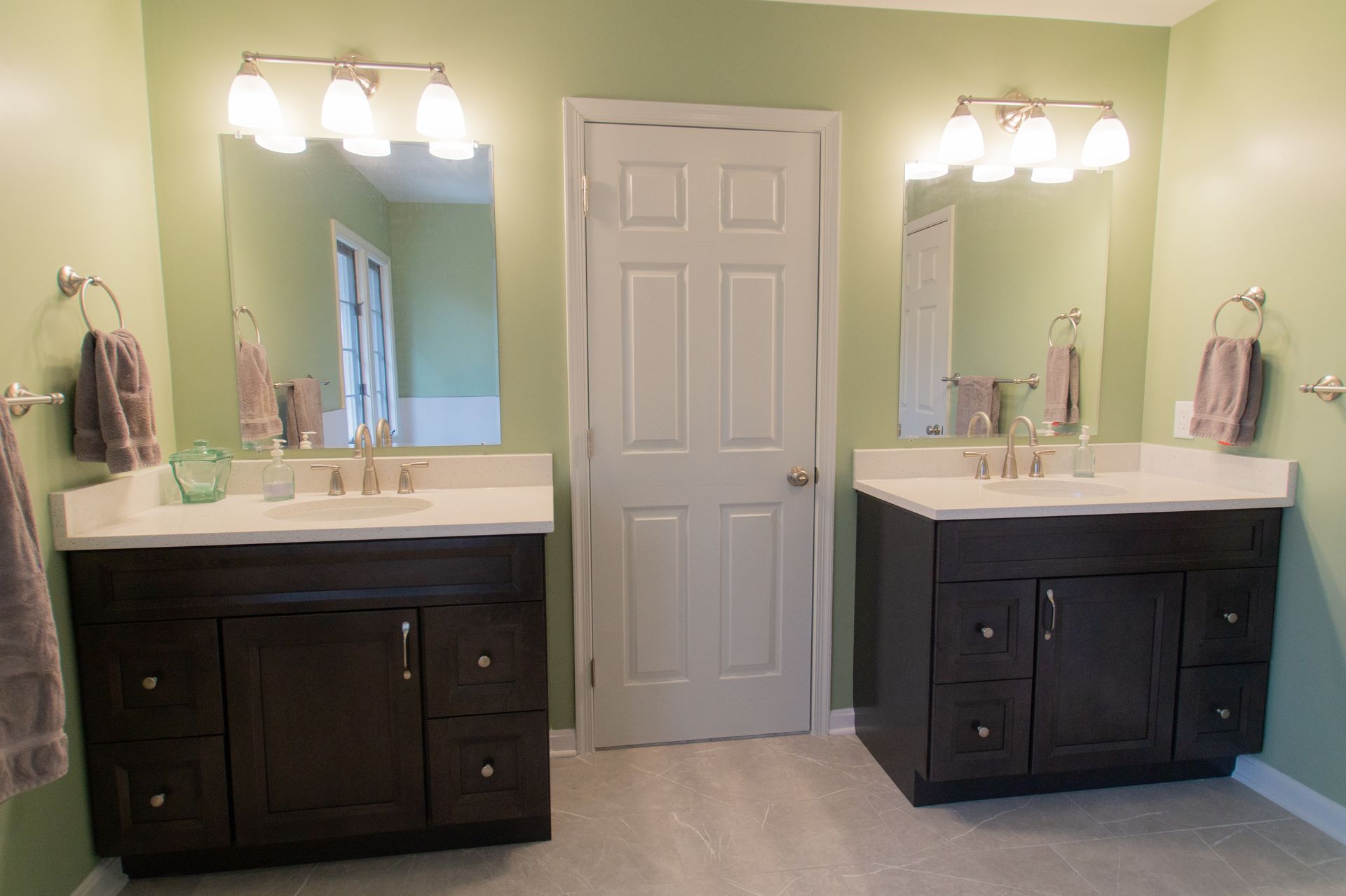 Two dark brown bathroom vanities with mirrors and sconces, flanking a white door, against a light green wall.