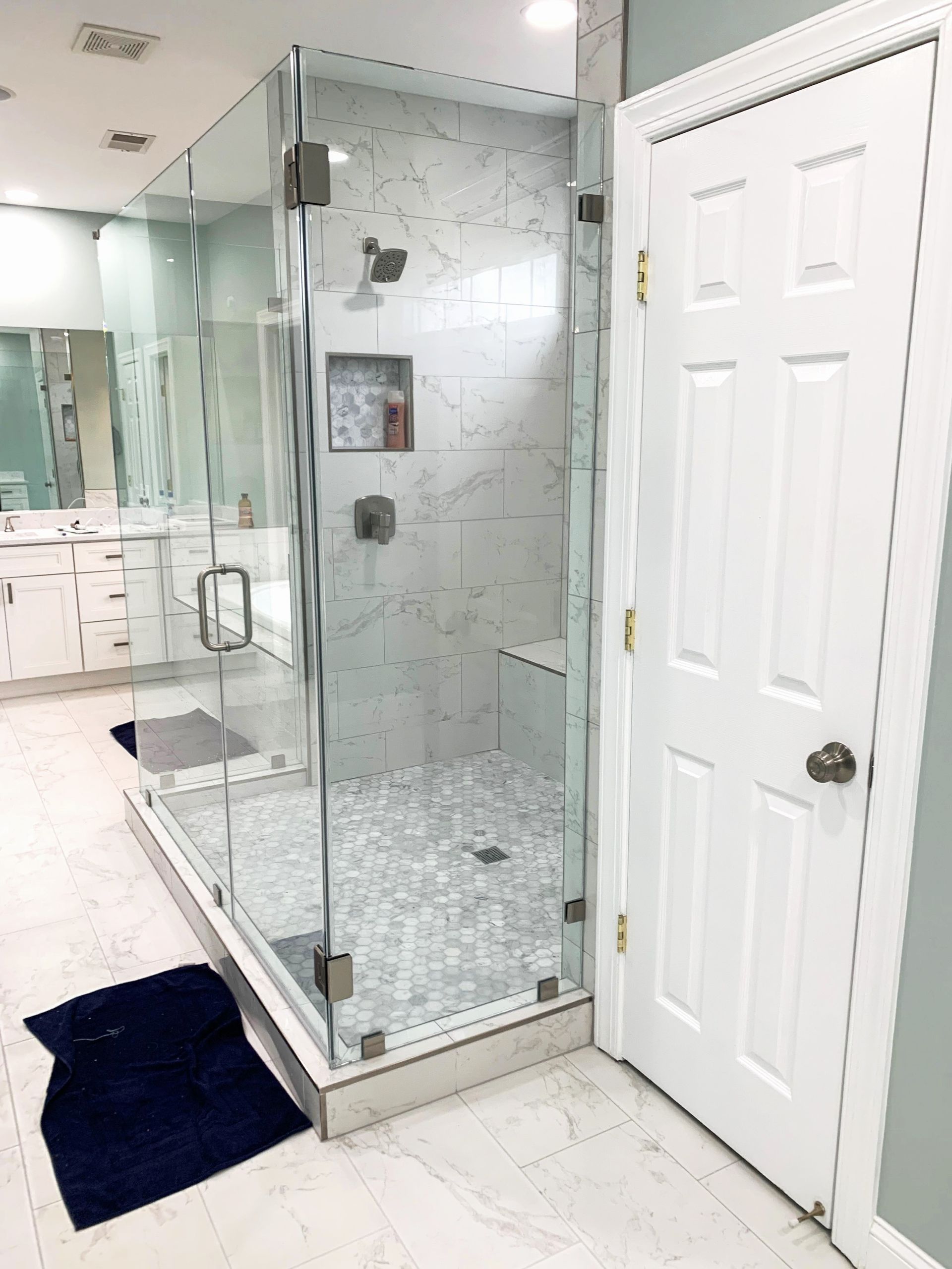 Spacious, modern bathroom with a glass shower enclosure, white marble tile, and a white door to the right.
