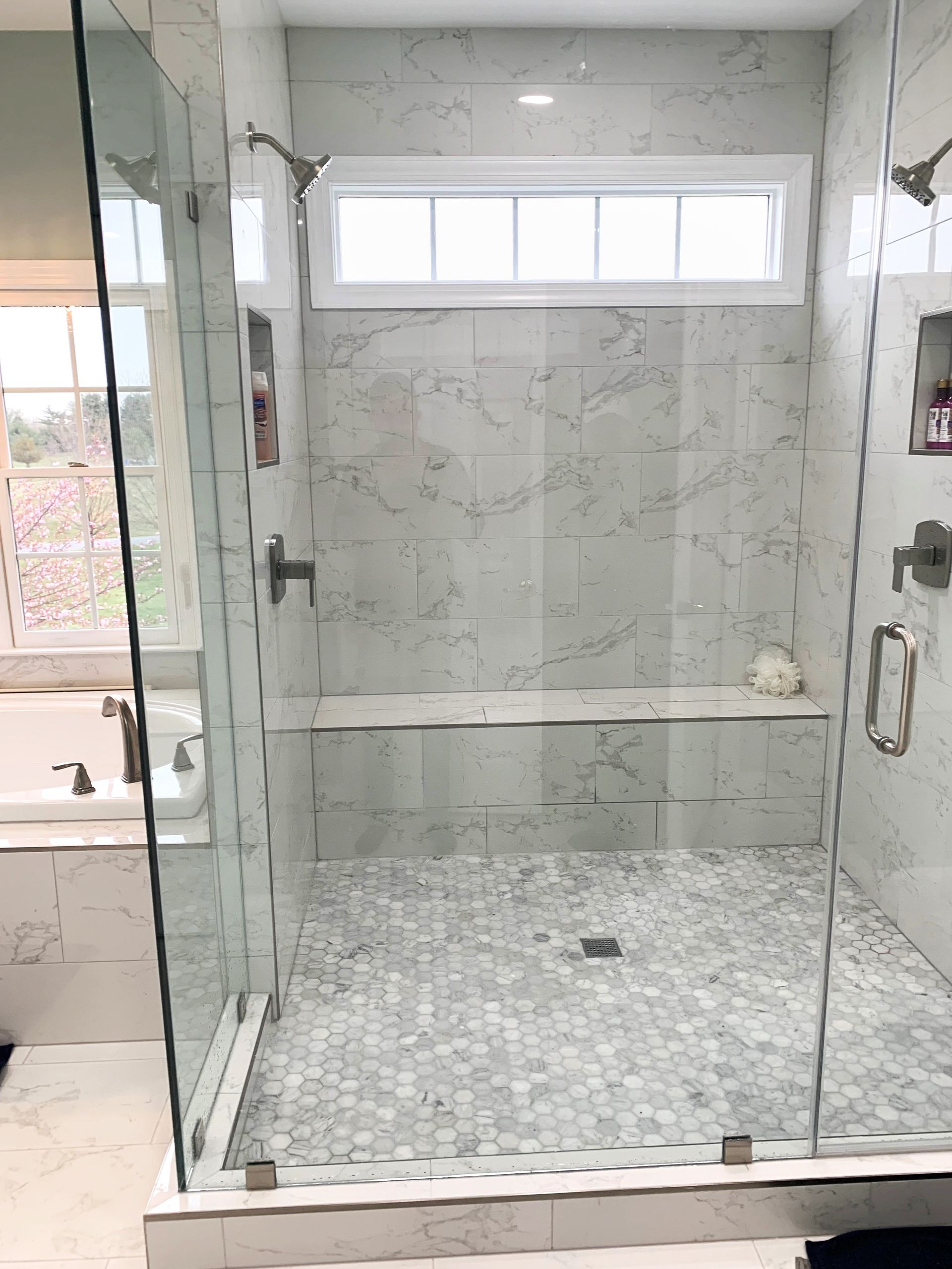 Modern, tiled shower with glass doors. Features marble-look walls, small mosaic floor, a built-in bench, and a window above.