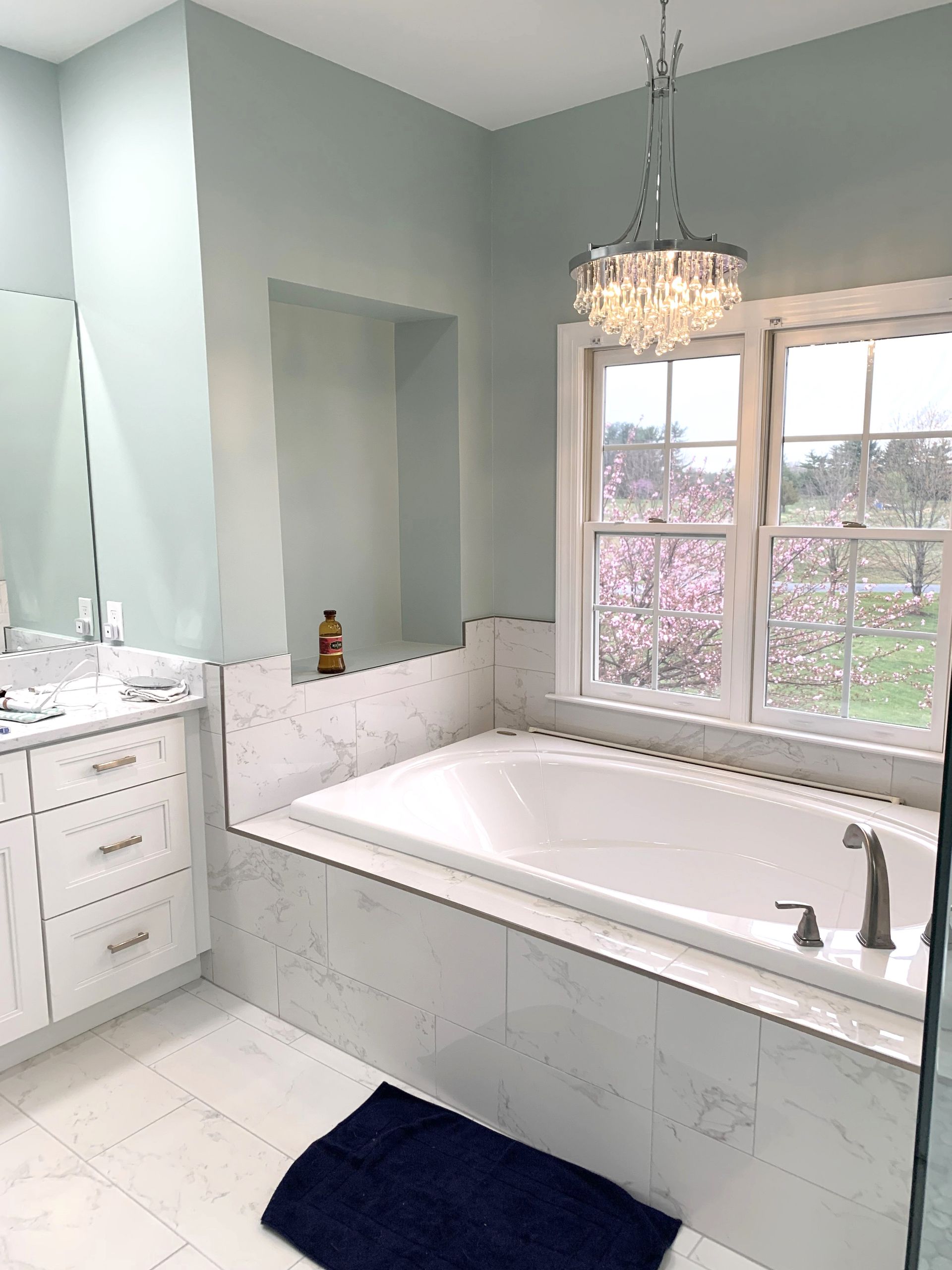A light blue bathroom with a white tub, marble tile, and a chandelier. A blue rug sits on the floor.