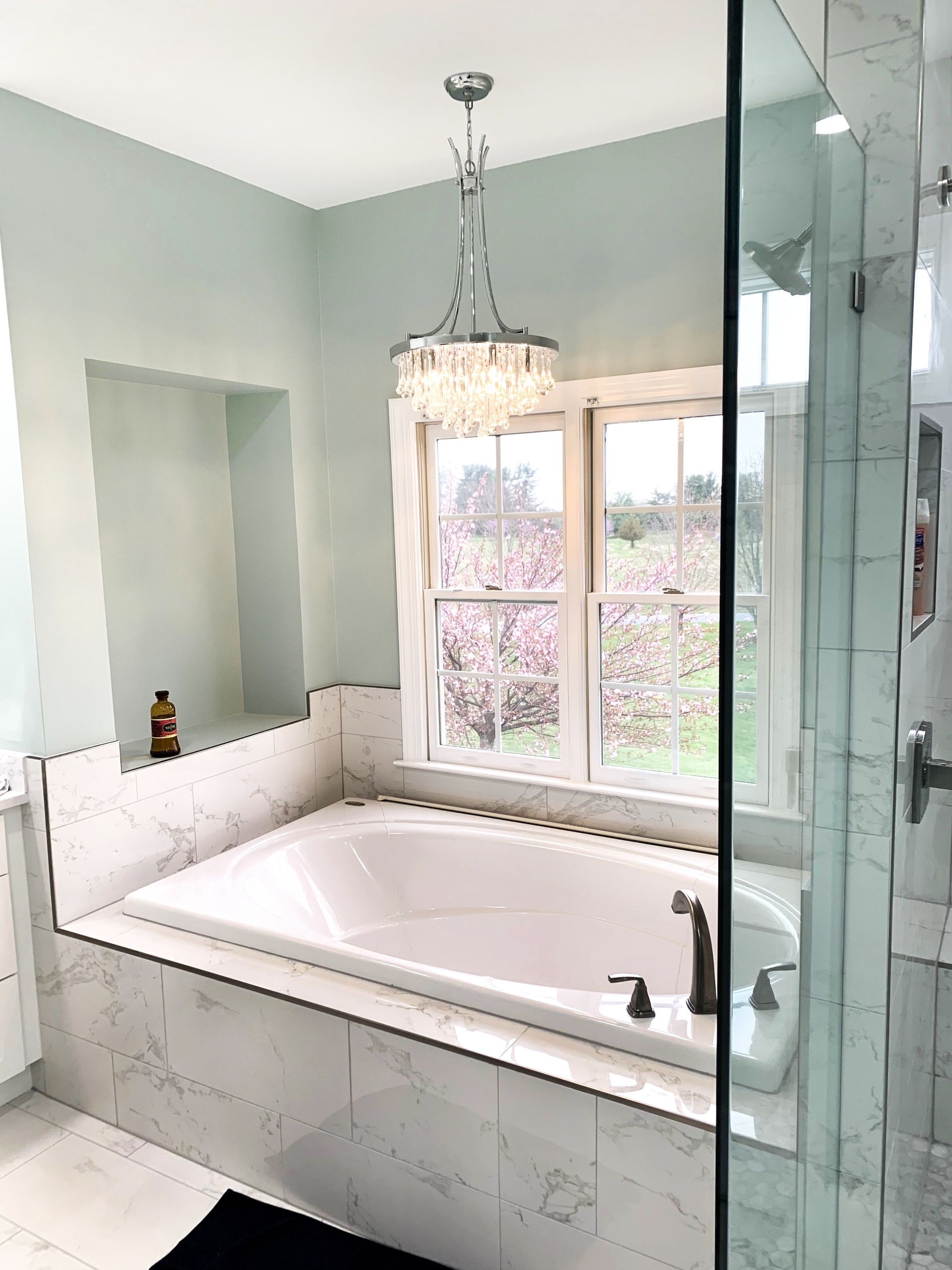 Elegant bathroom with a white bathtub, windows overlooking a garden, and a crystal chandelier. Glass shower door is on the right.