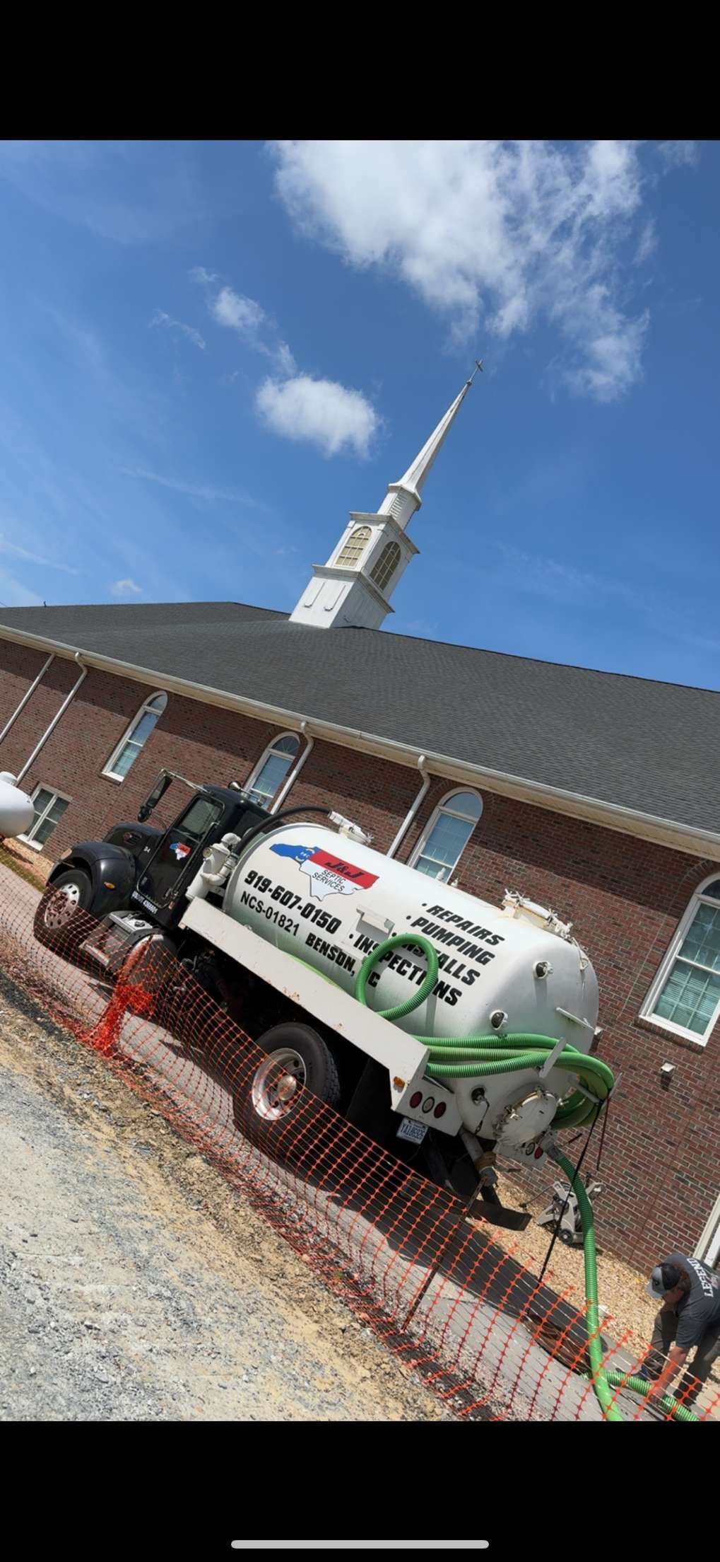 A septic tank truck is stuck leaning against a brick church. Clear blue sky with a white steeple.