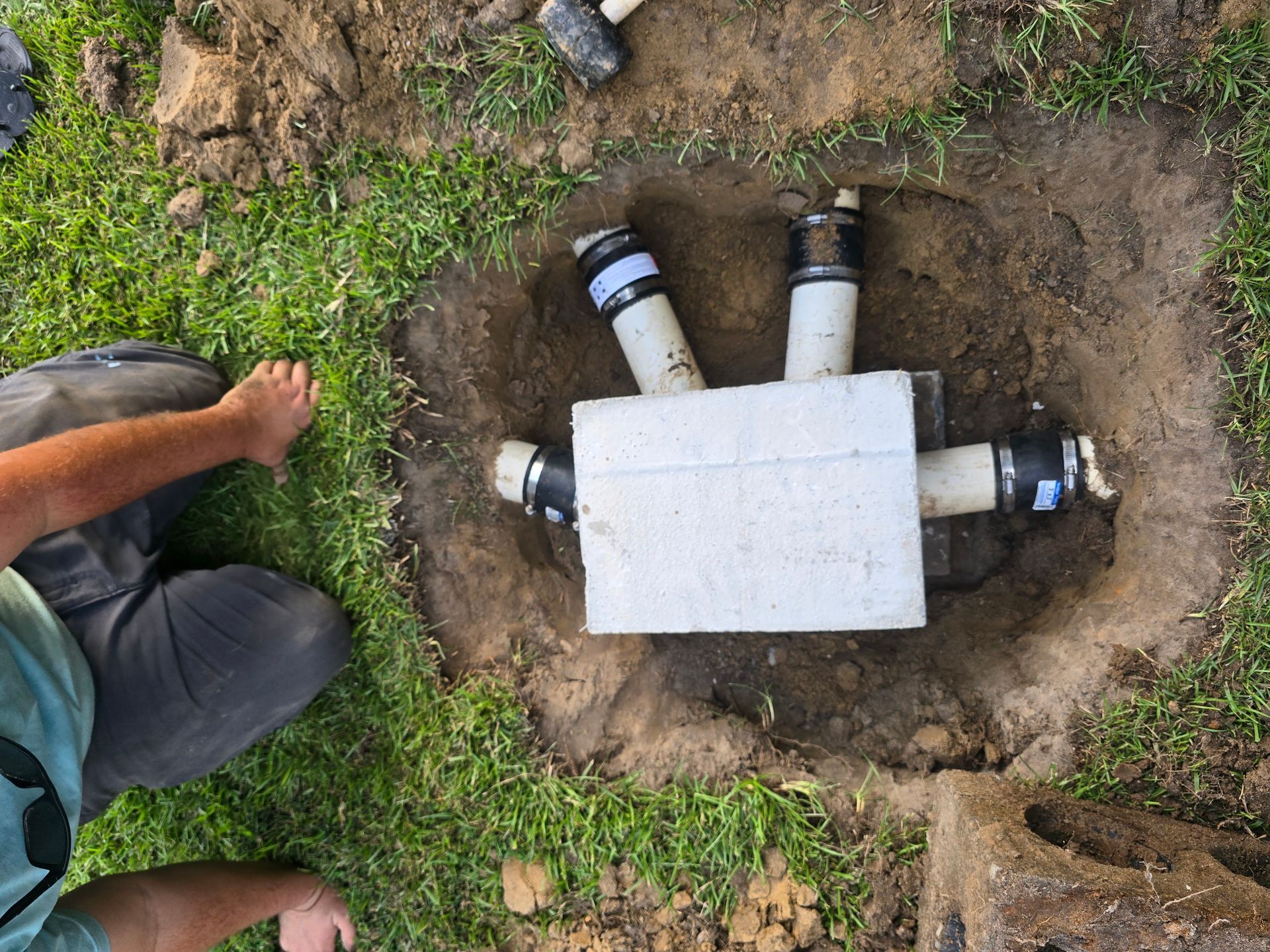 Man working on buried white box with connected pipes in grass.