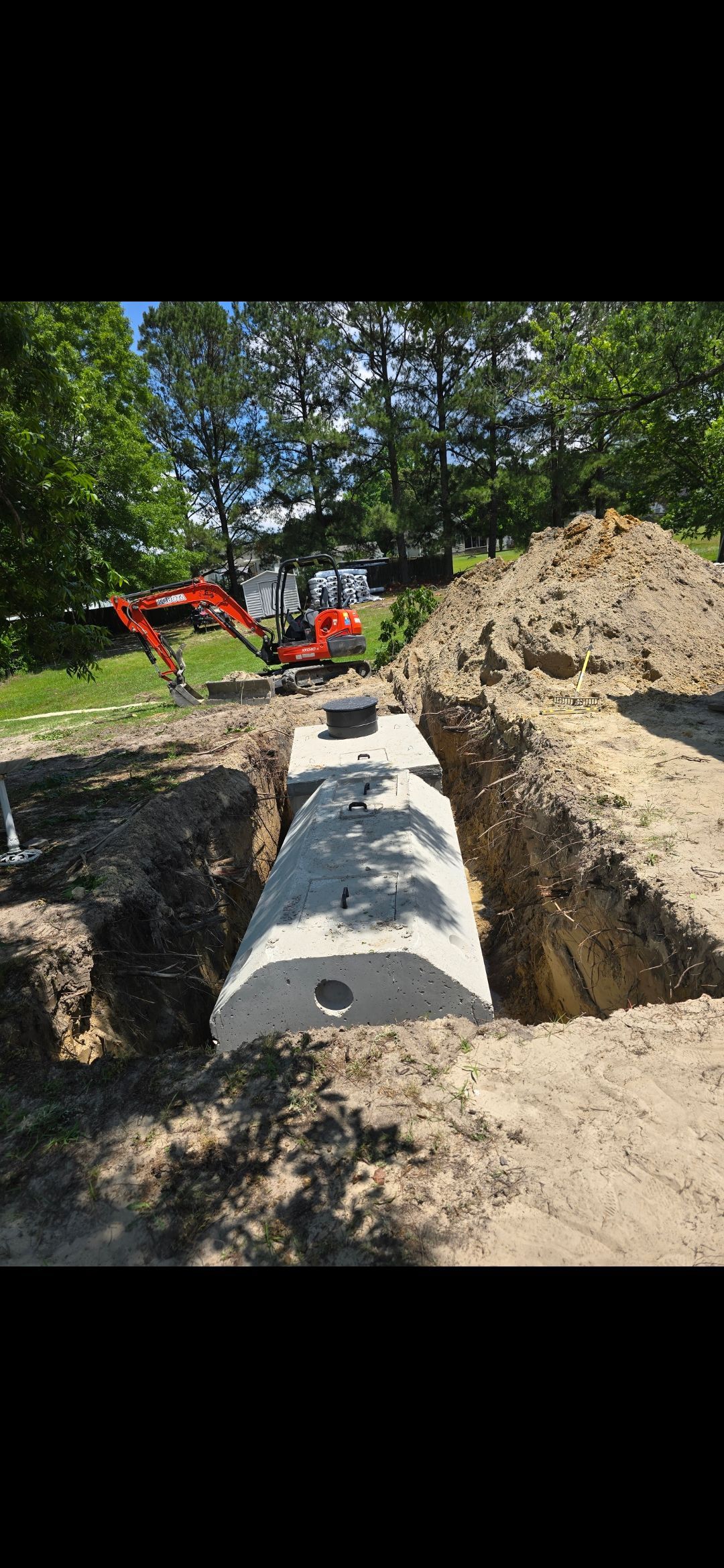 A concrete septic tank in an excavated trench, with an excavator in the background.