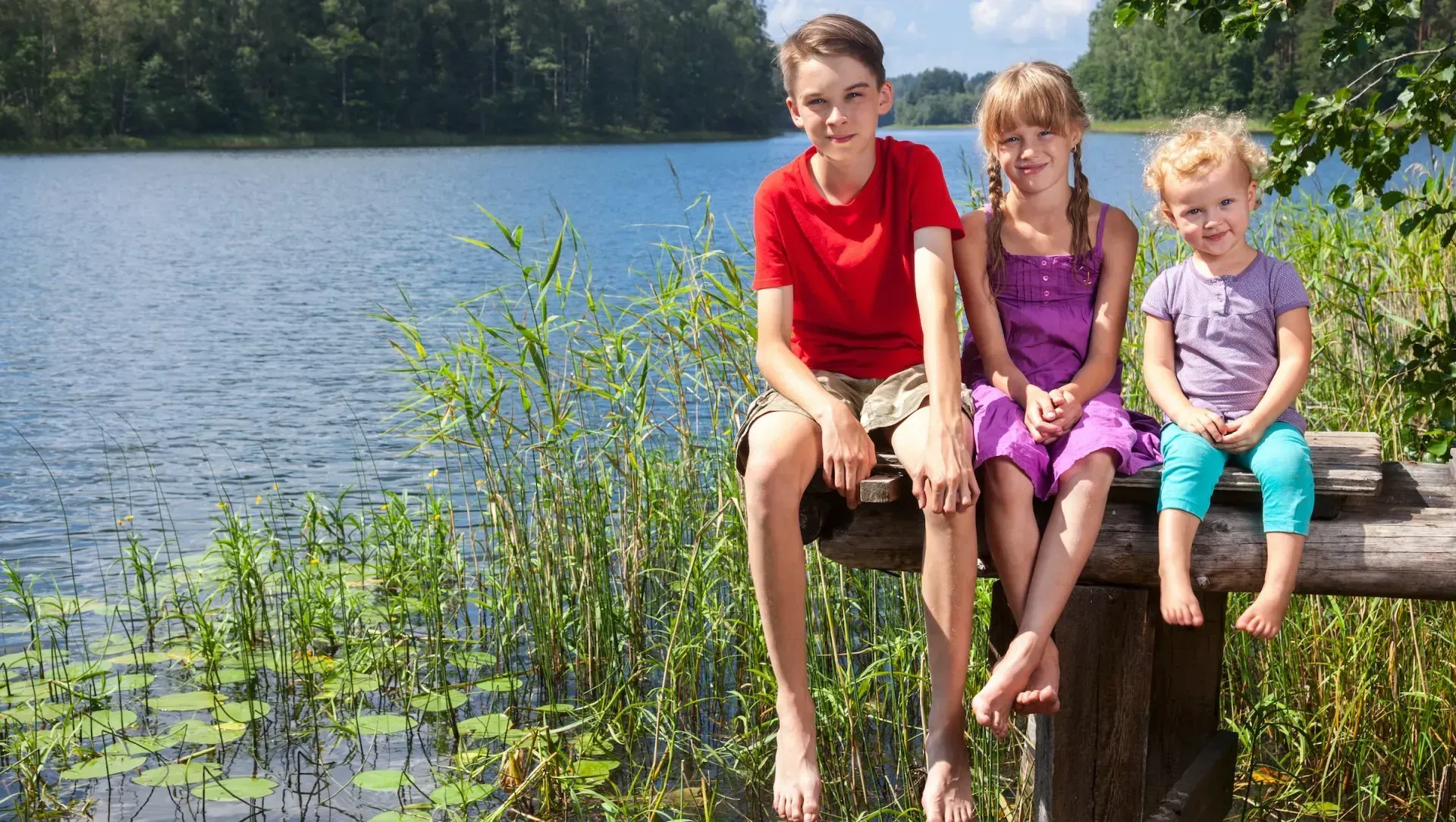 Three Montessori students are sitting on a wooden dock near a lake.