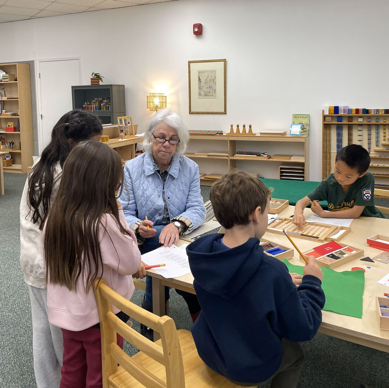 A Montessori teacher works with elementary students at a table using various math materials including bead bars and a checkerboard.