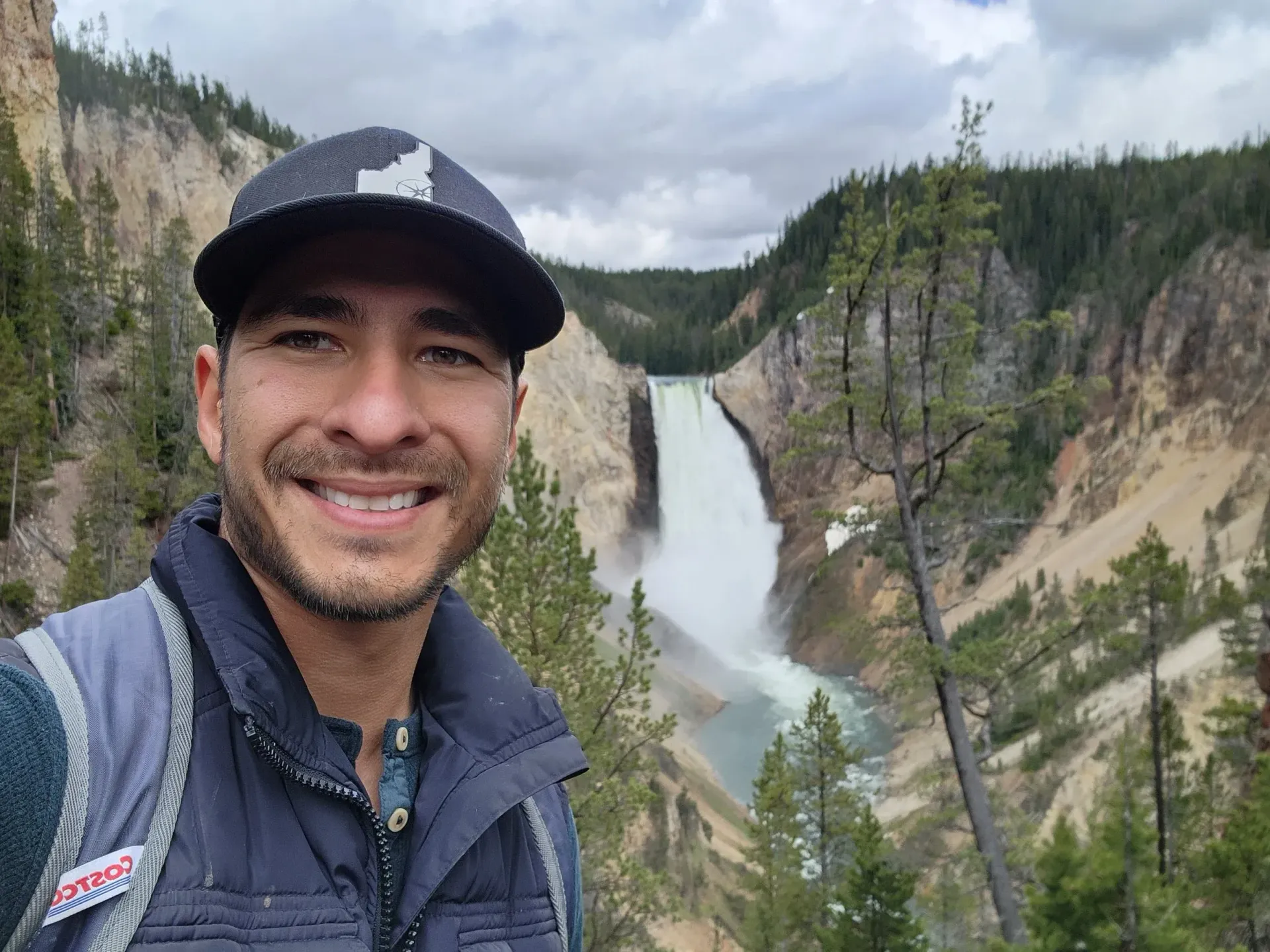 Man smiles at camera, standing before Lower Falls in Yellowstone National Park.