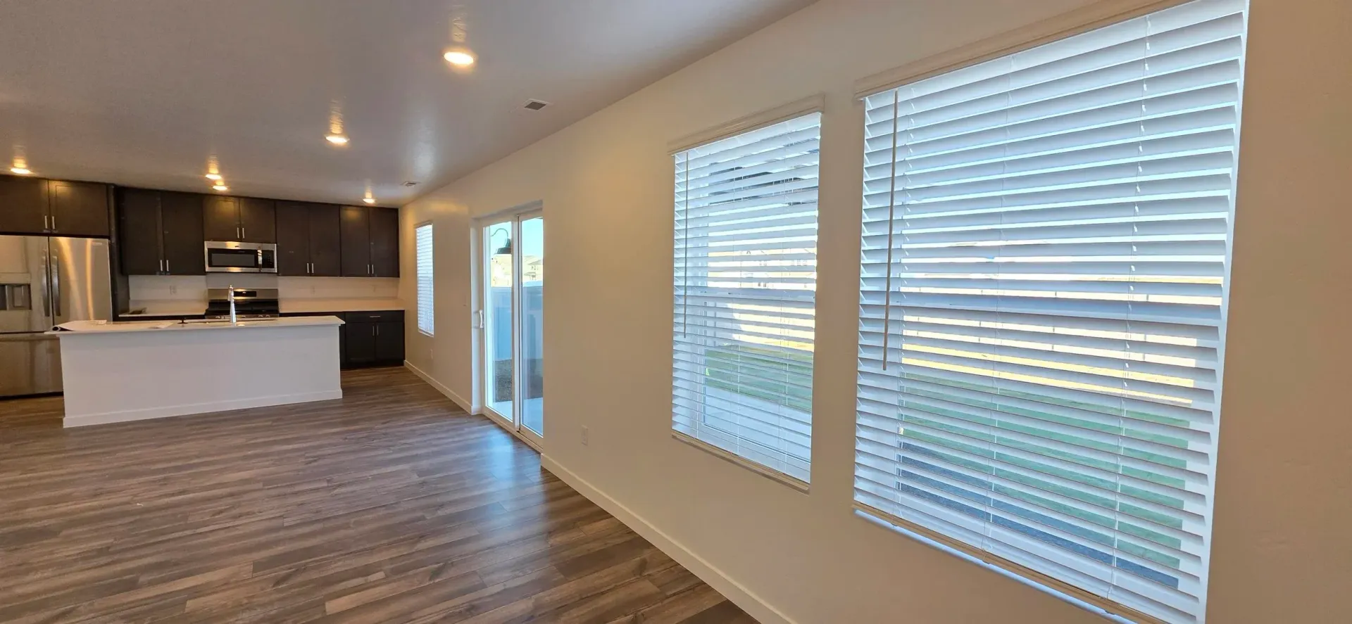 Interior of a modern kitchen and living space with dark cabinets, a white island, and large windows with blinds.