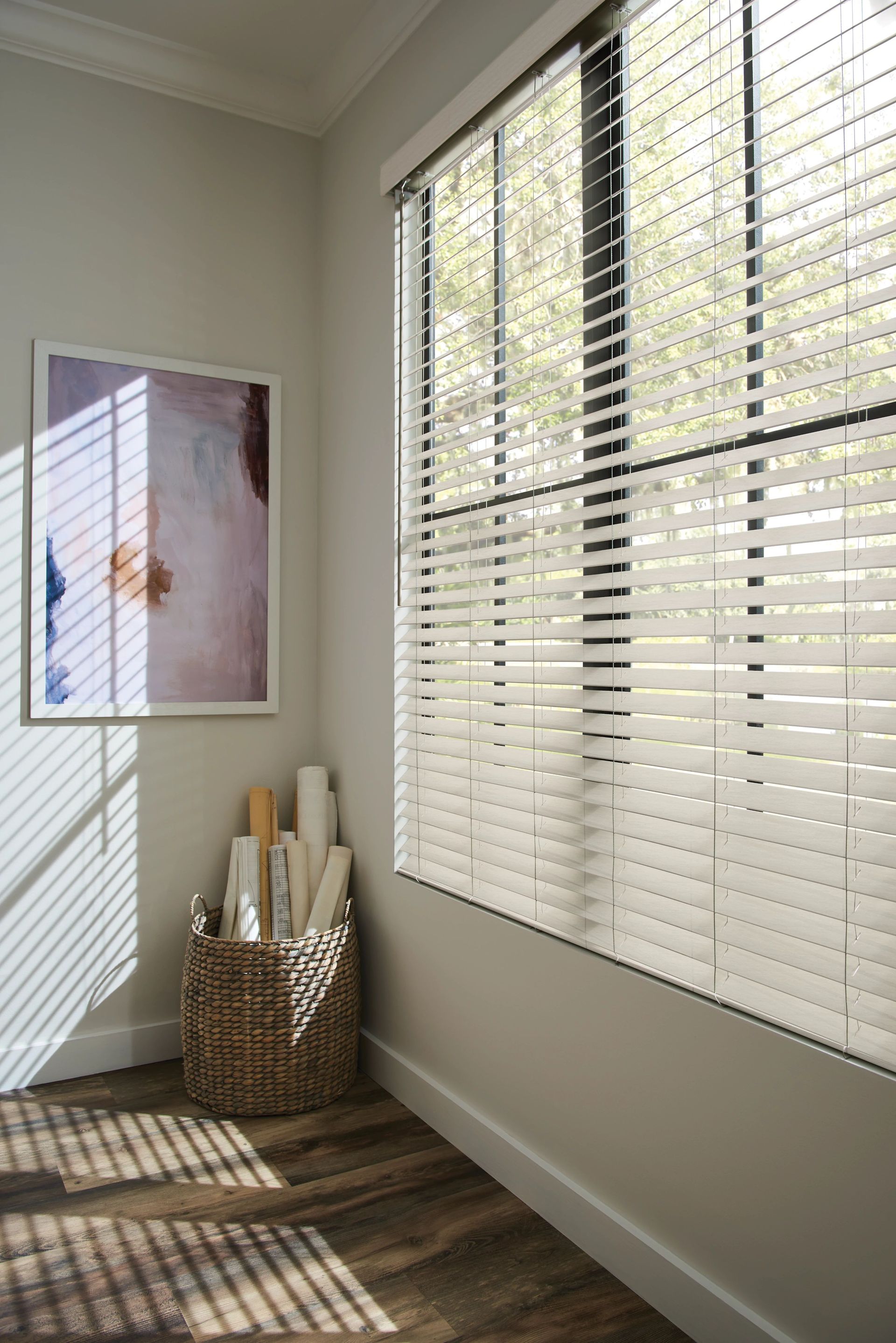 Room with window blinds, artwork, and basket of rolls. Sunlight streams across wooden floor.