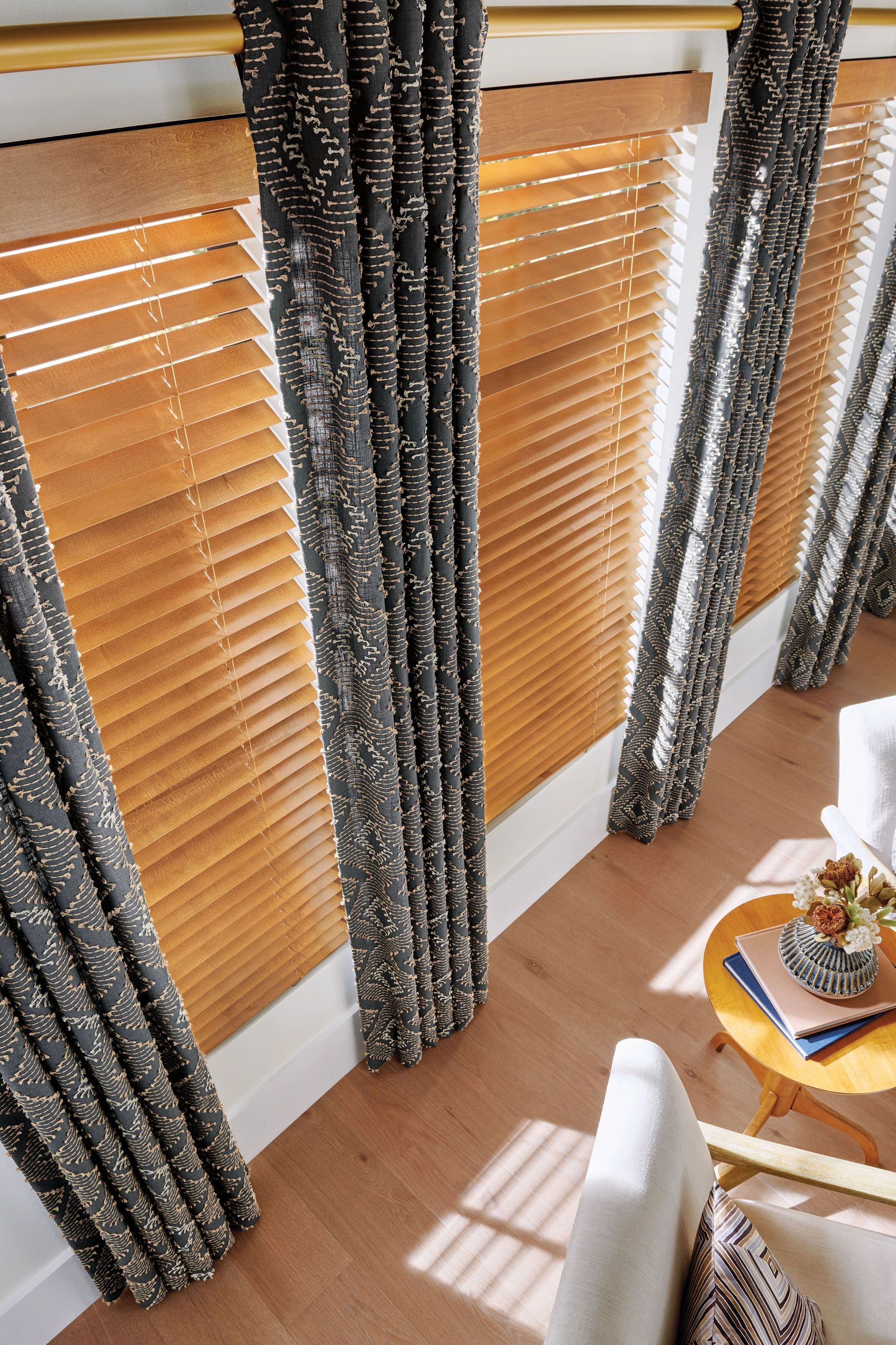 Wooden blinds and patterned curtains in a room with light wood floors and a small side table.
