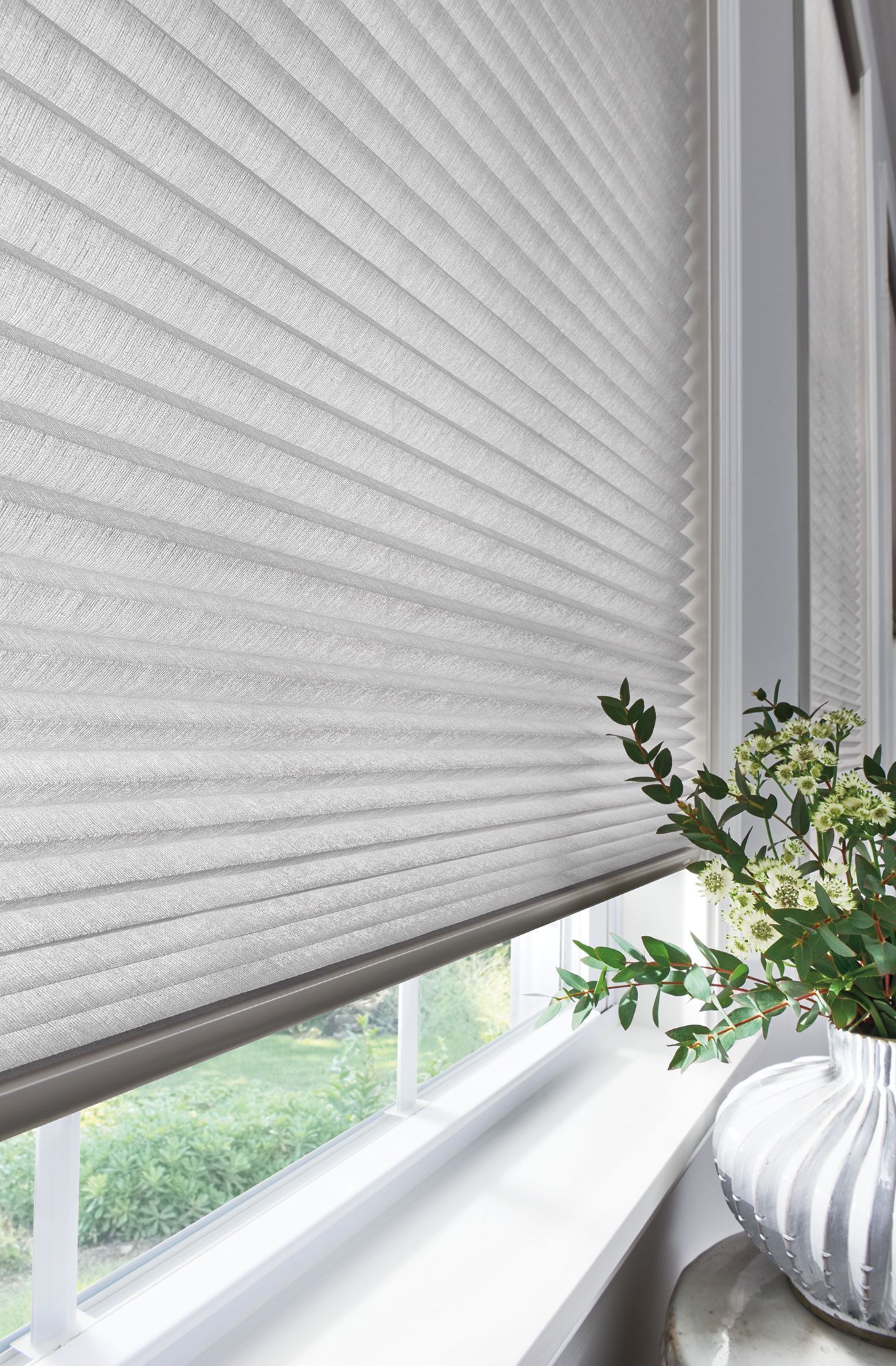 Light-colored pleated window blinds cover a window; a vase with greenery sits on the windowsill.