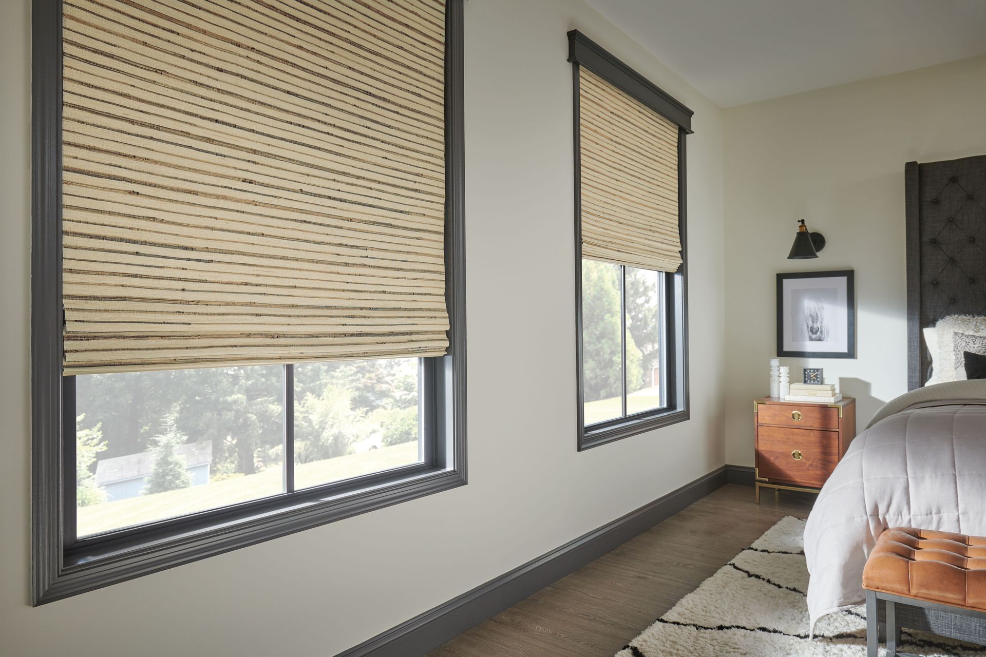 Two windows with woven blinds in a room with a bed and wood flooring.