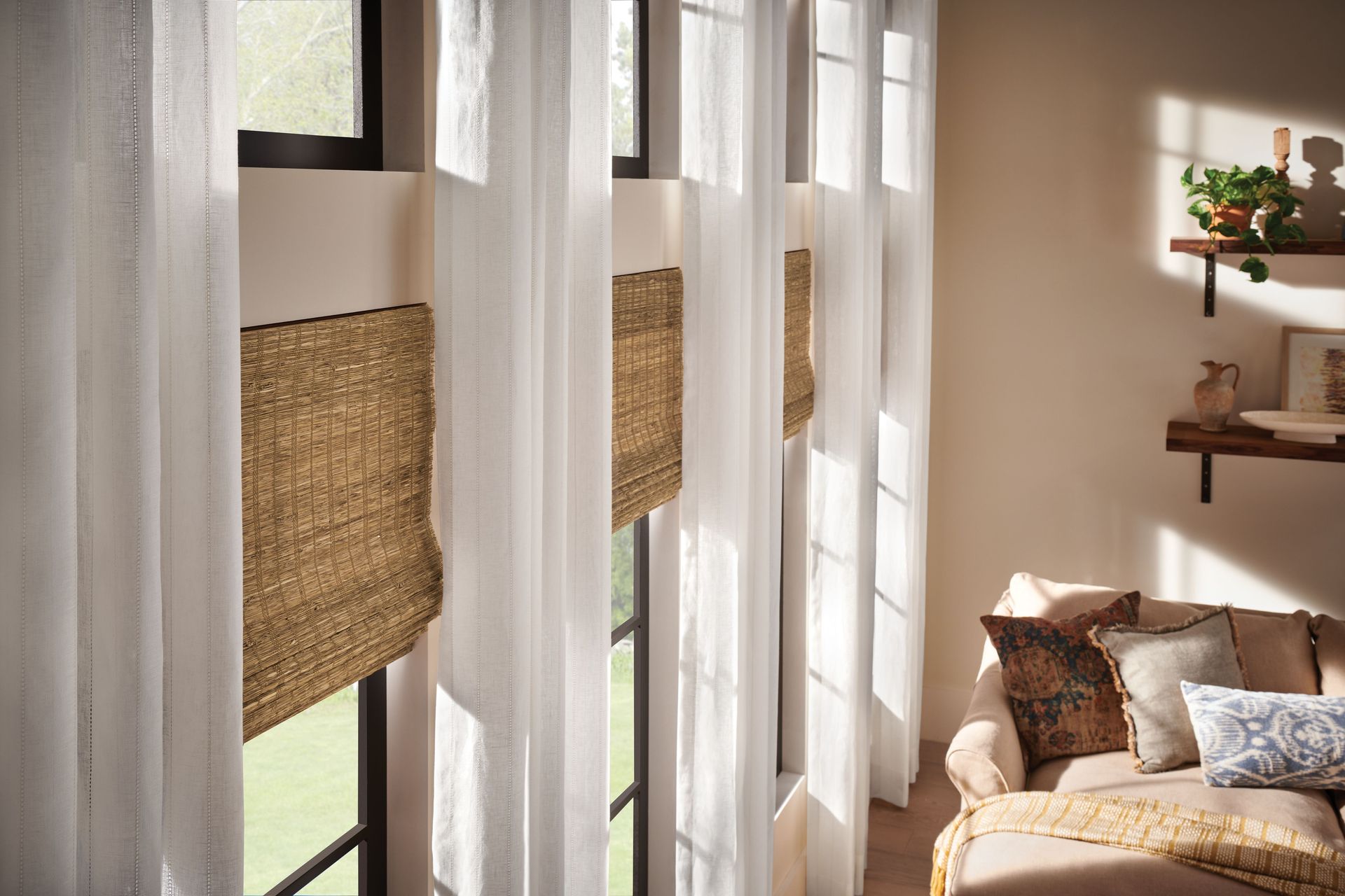 Sheer white curtains and woven blinds frame windows; a sofa and shelves are in the room.
