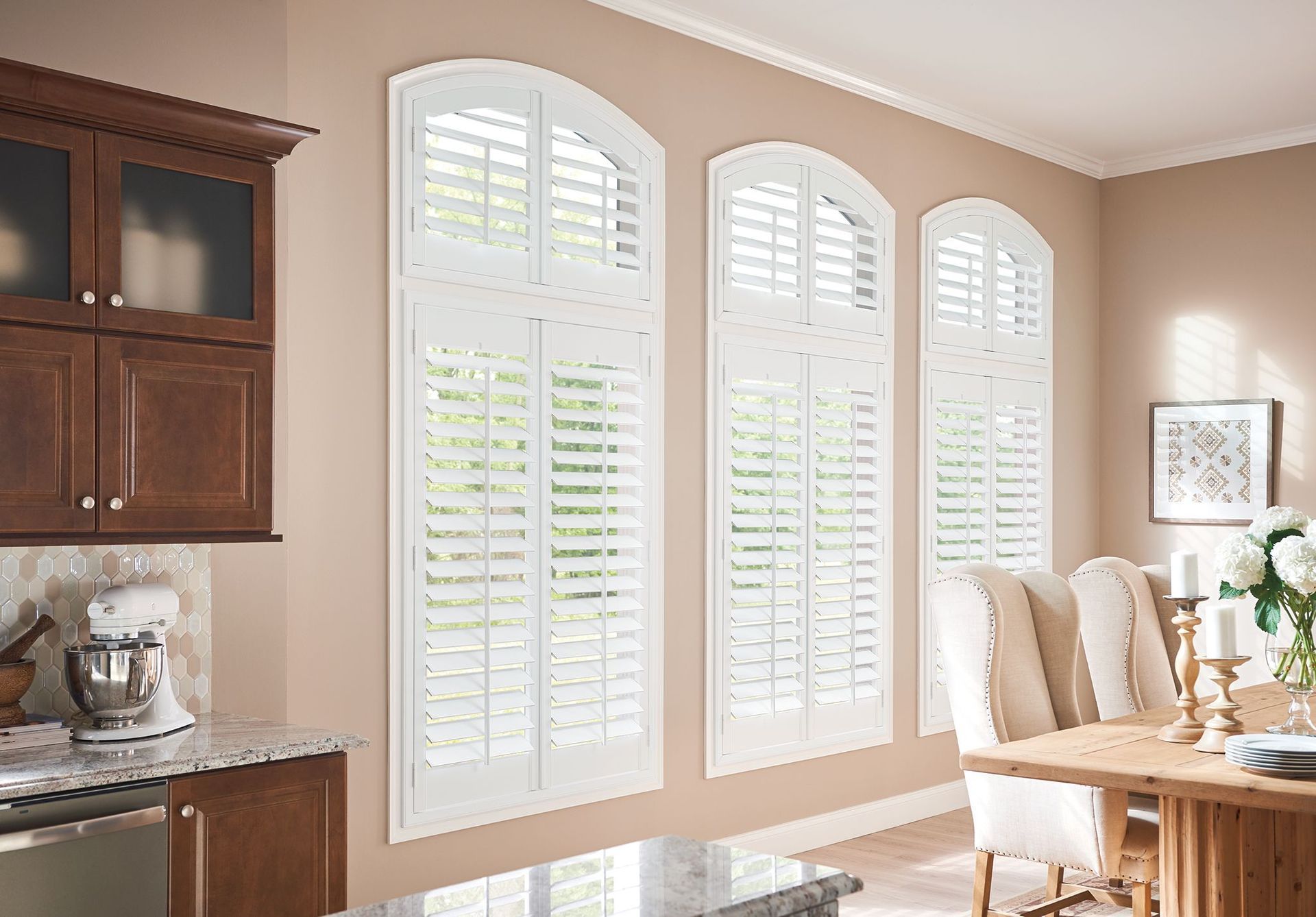 Three arched windows with white shutters in a neutral-toned room; dark wood cabinetry to the left, dining table to the right.