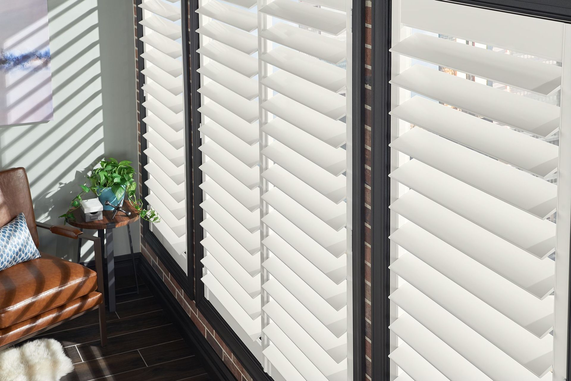 White shutters with angled slats cover a window, letting in striped sunlight. A leather armchair sits nearby.