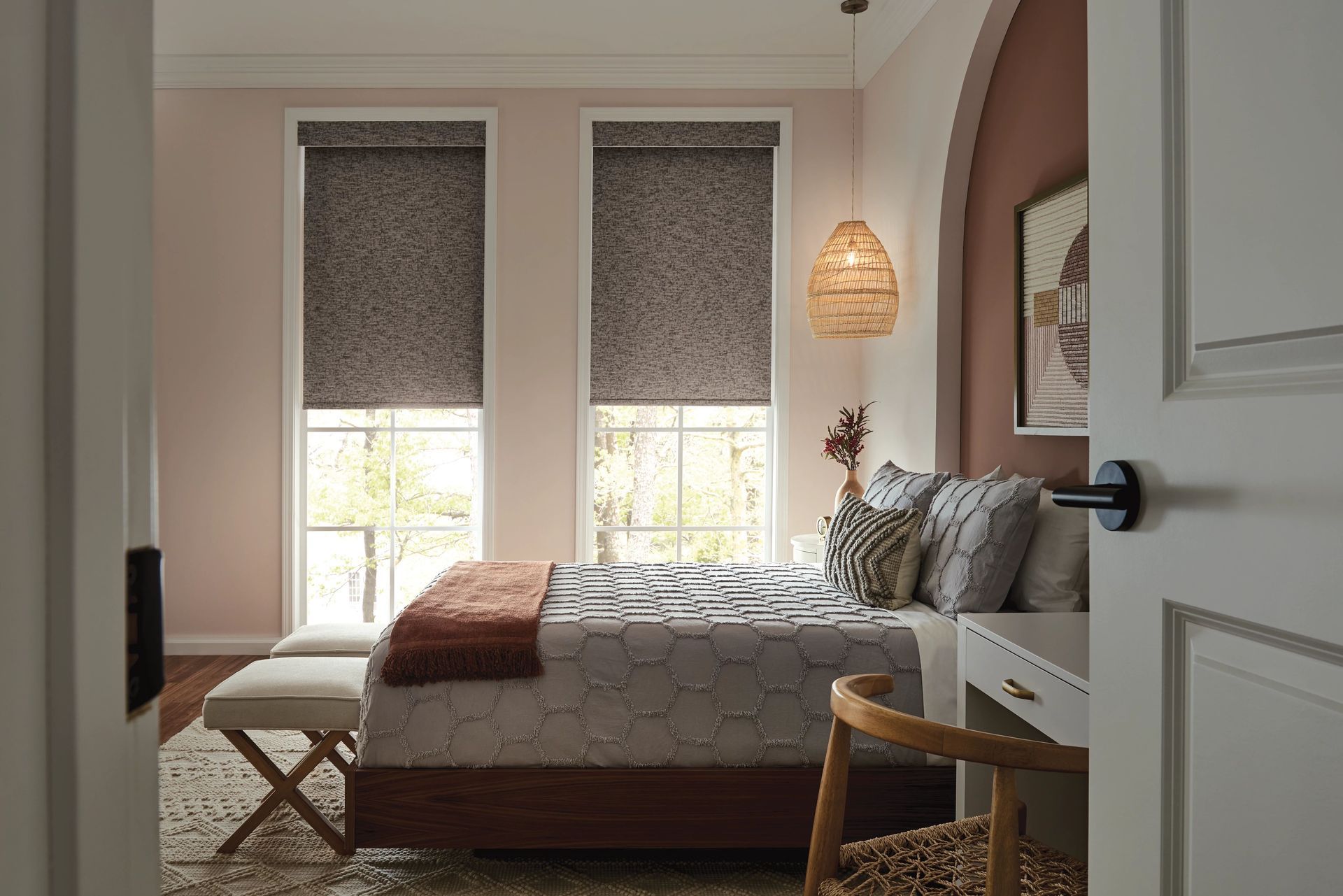 Bedroom with two windows, bed, desk. Dusty rose walls, patterned blinds, neutral bedding, woven lamp.