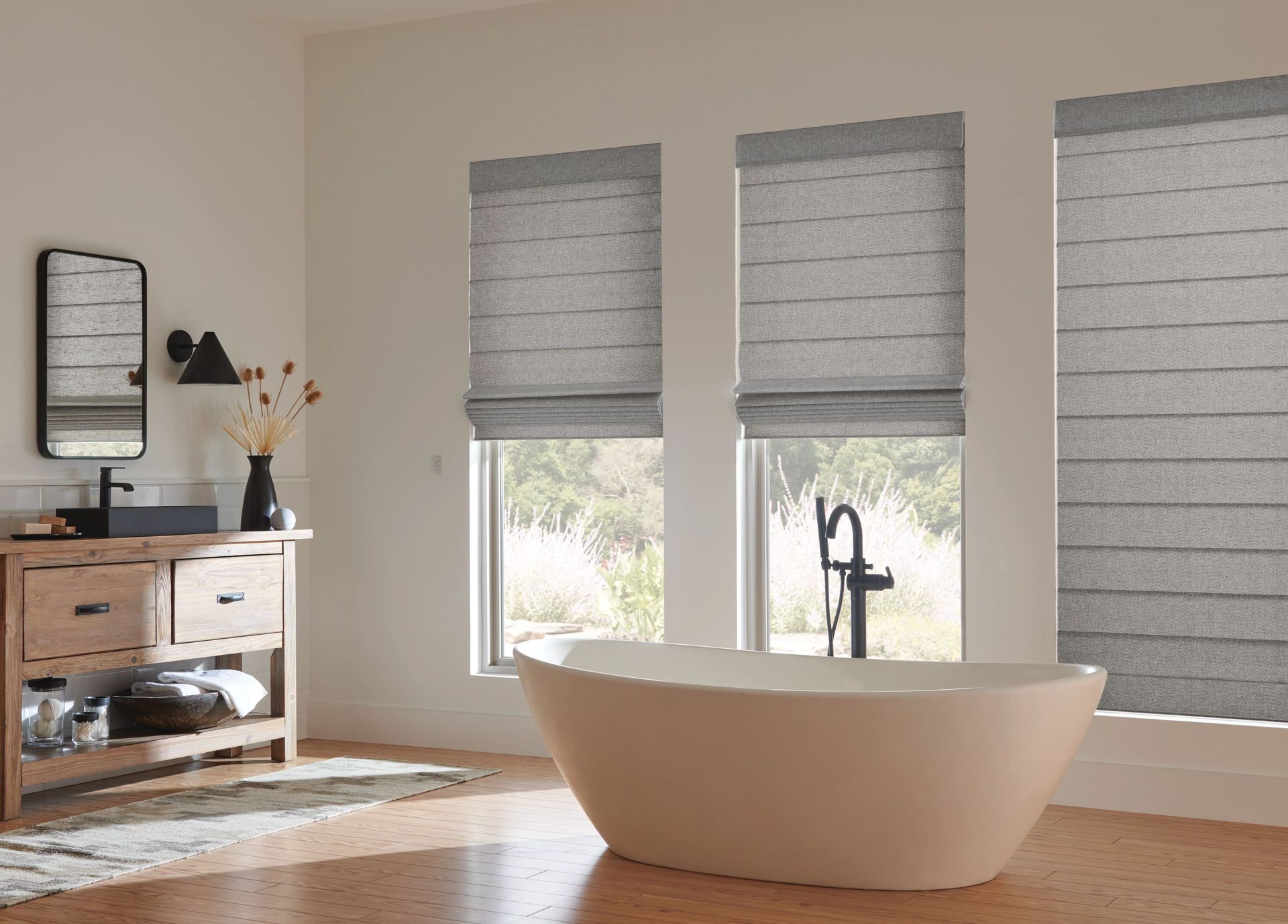 Bathroom with white soaking tub, wooden vanity, and windows with gray Roman shades.