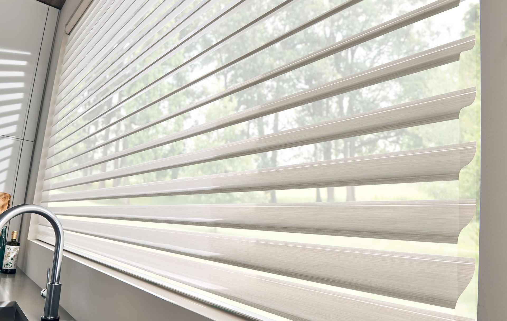Window with layered, light-colored blinds partially open, allowing filtered light and a view of trees.