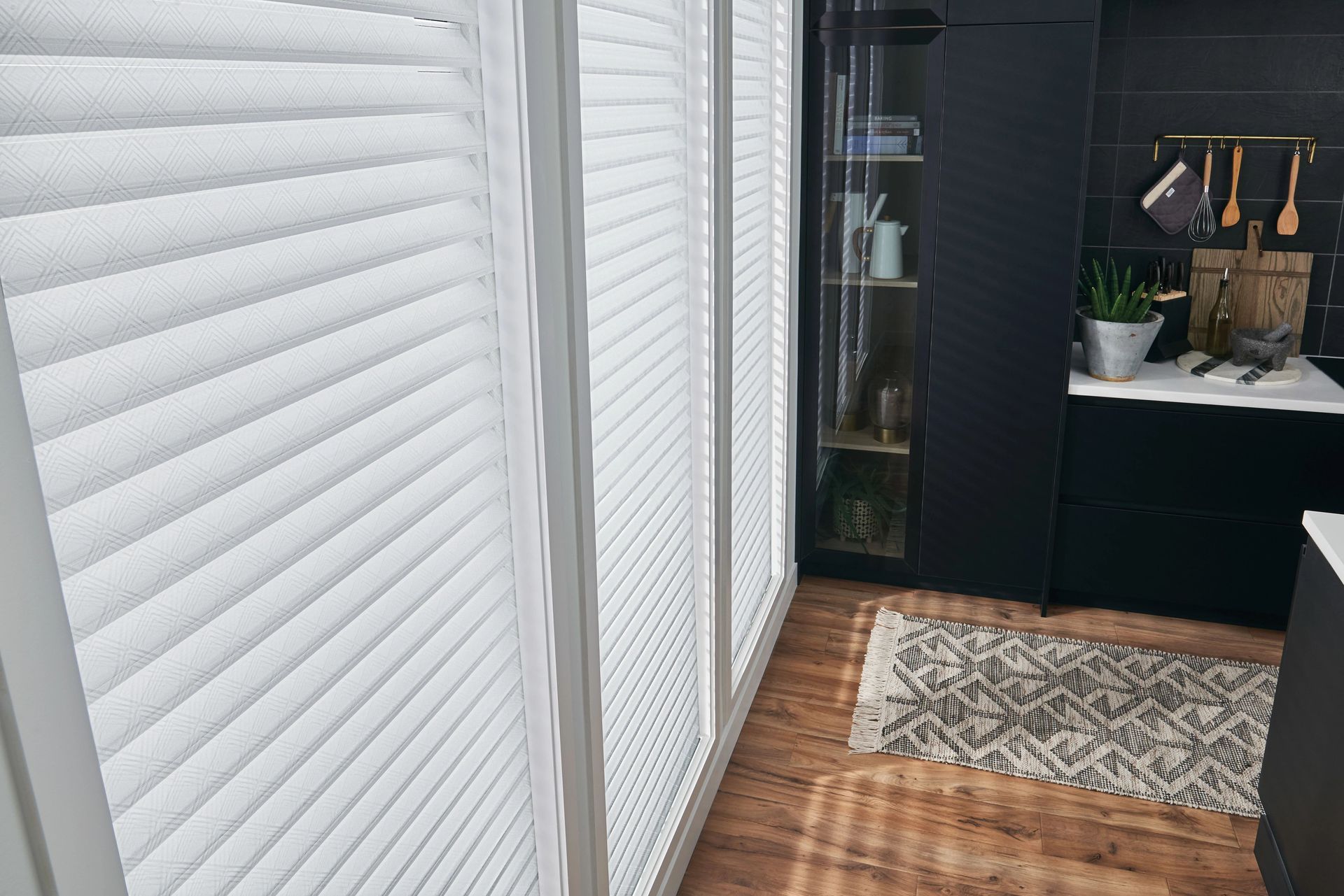 White pleated blinds in a kitchen, reflecting light. Dark cabinets and wood floor are visible.