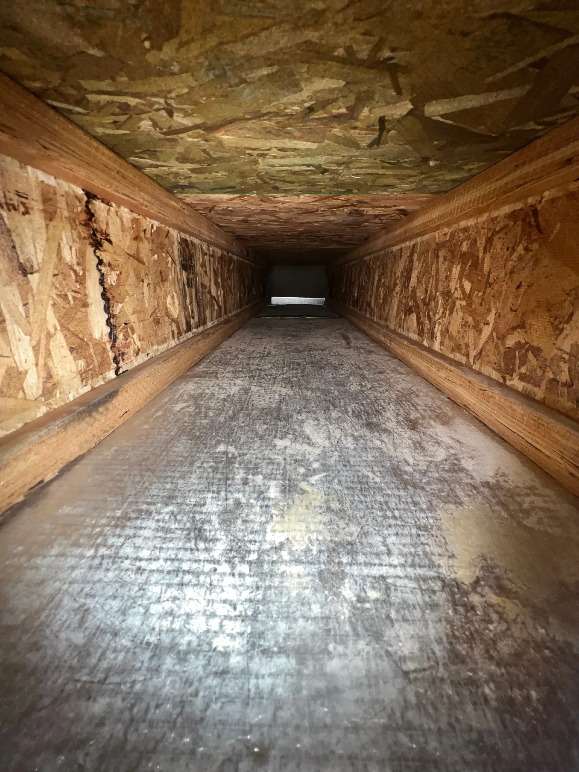 Long, narrow wooden tunnel with a rough floor and a bright opening at the far end