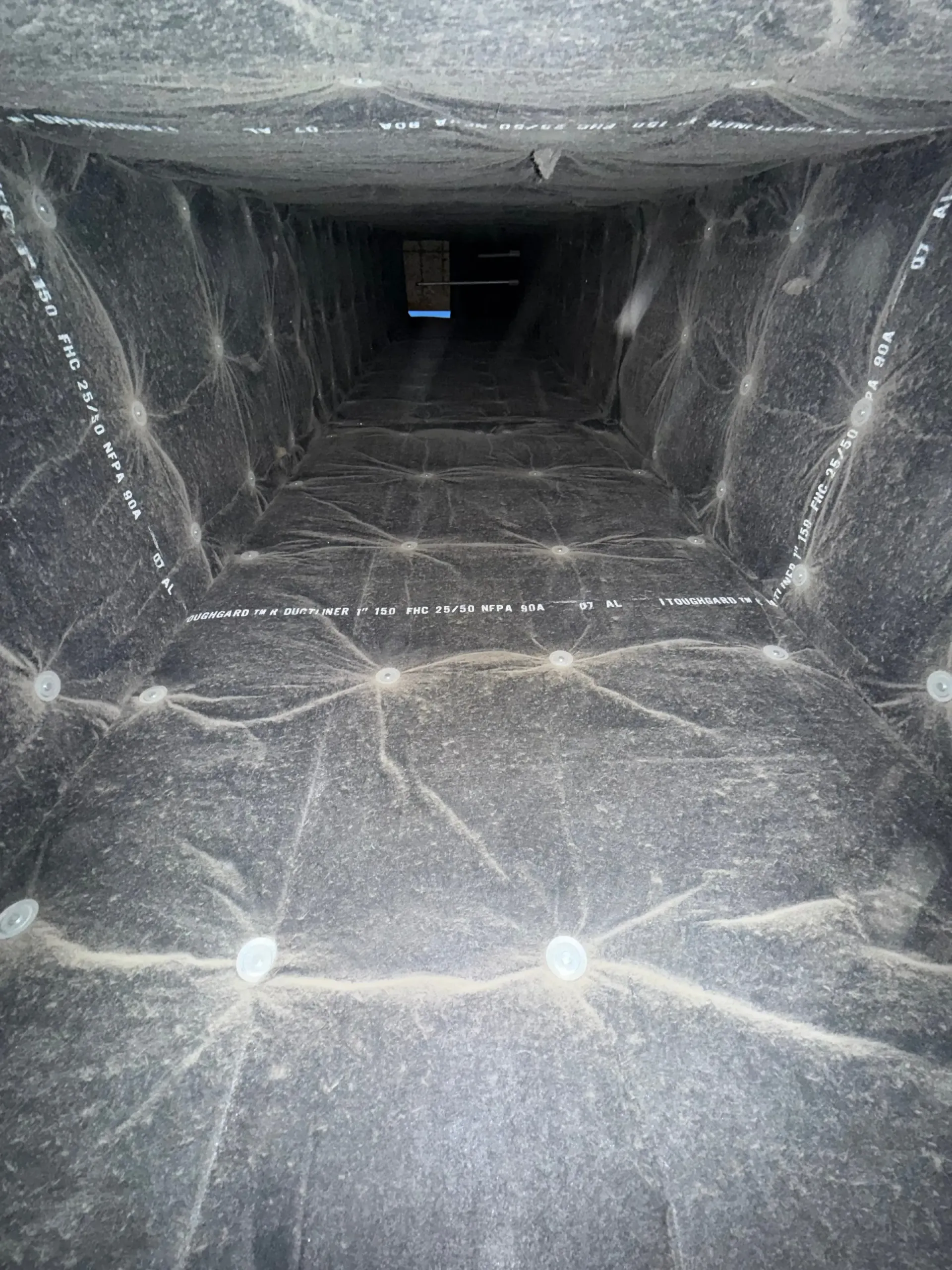 Interior of a dusty metal ventilation duct with lights near the foreground and a dark opening ahead