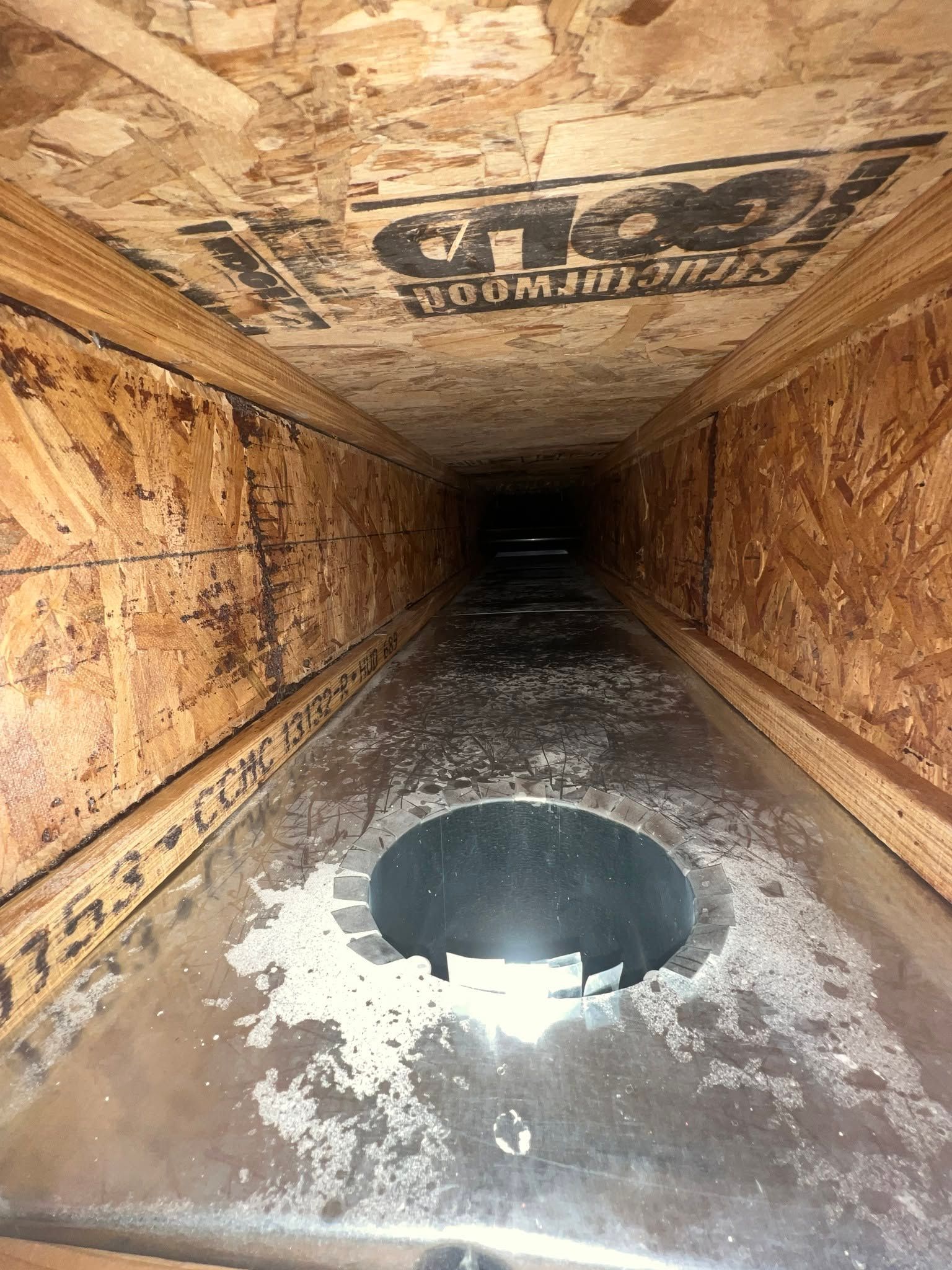 Damaged wooden crawlspace with a large round hole in the floor and a dark tunnel-like interior.