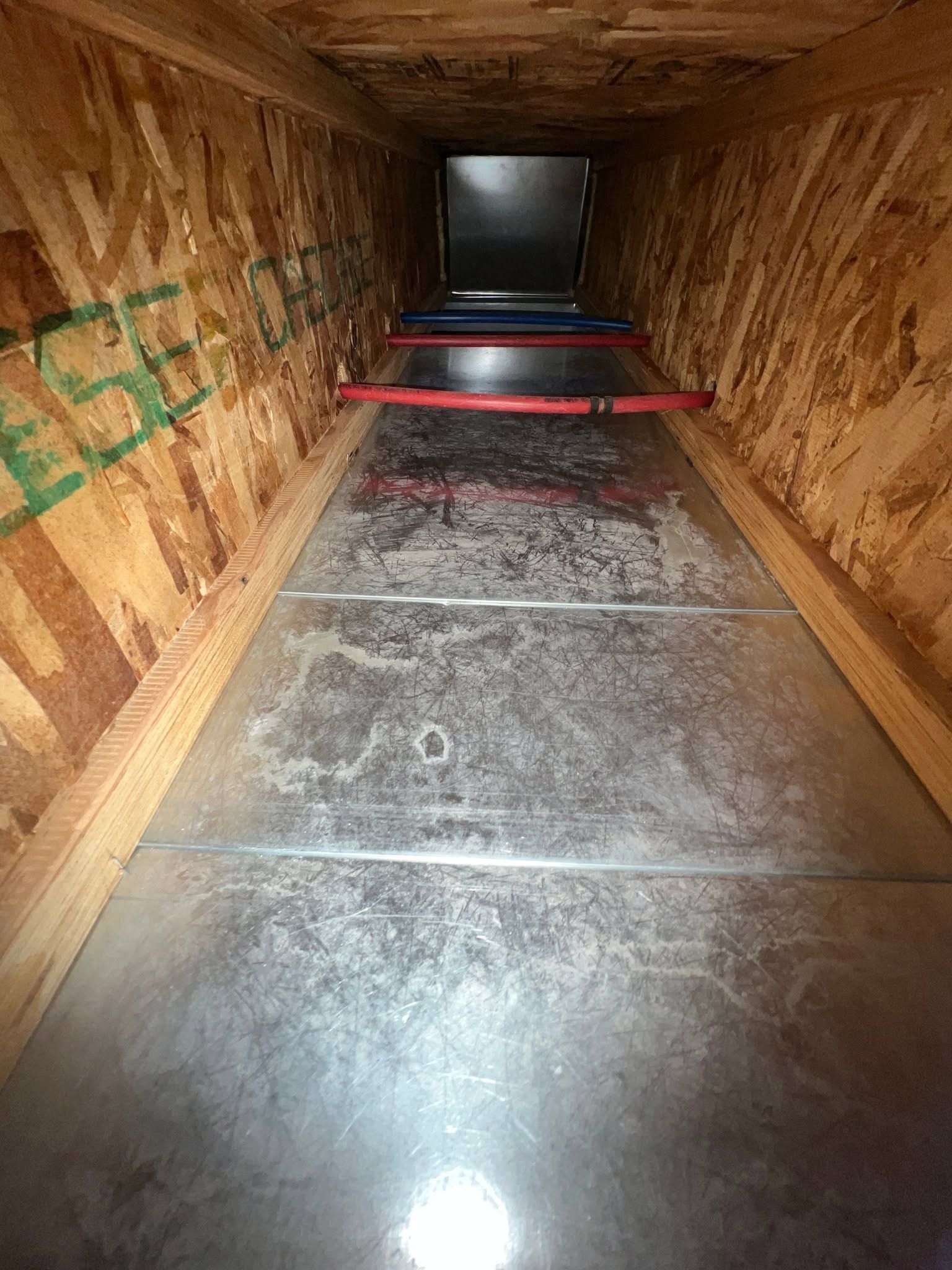 Inside a wooden cargo container with a metal floor and red bars across the opening, viewed from the entrance