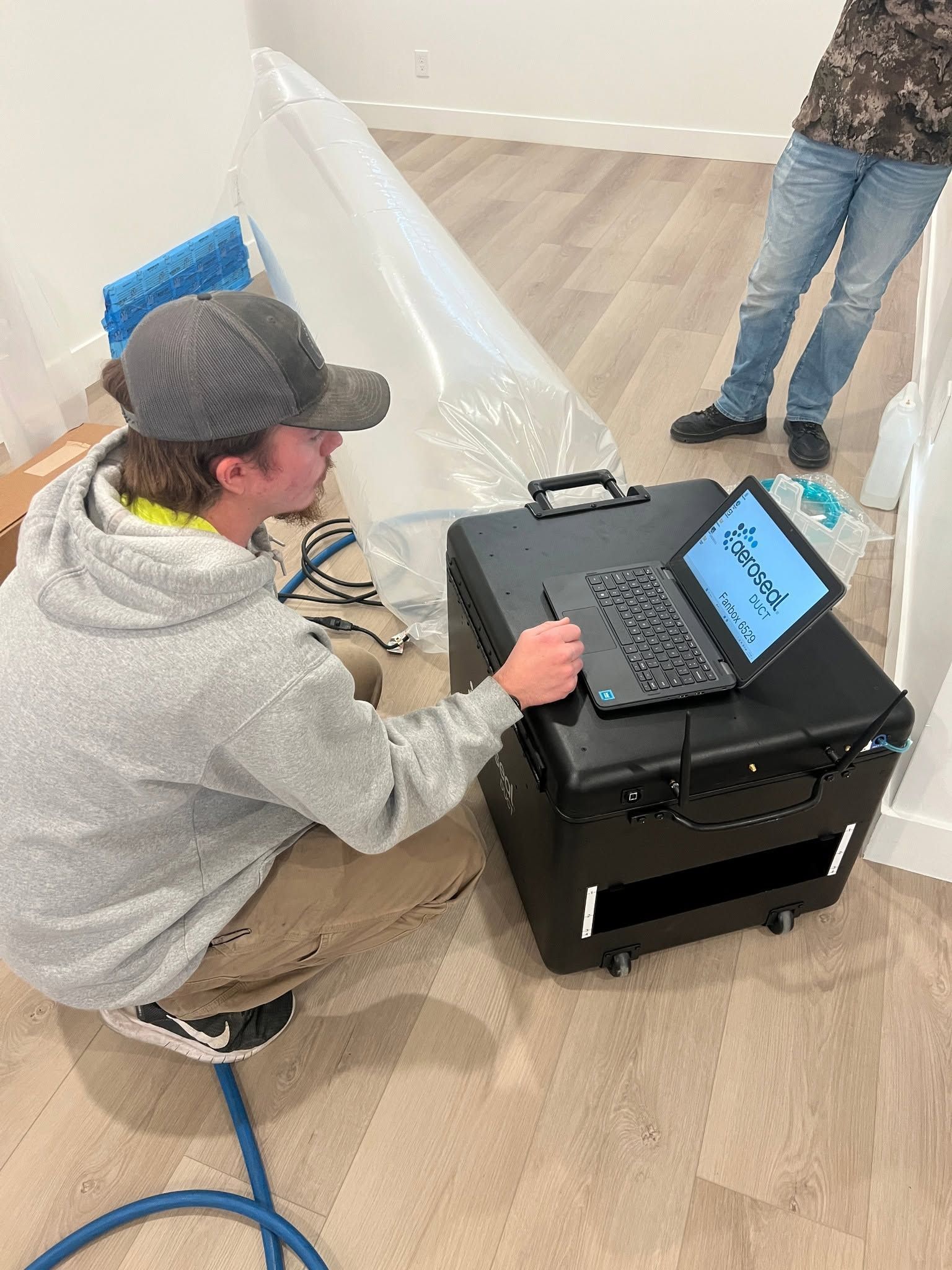 Person crouches beside a black device, using a laptop in a room with a wood floor.