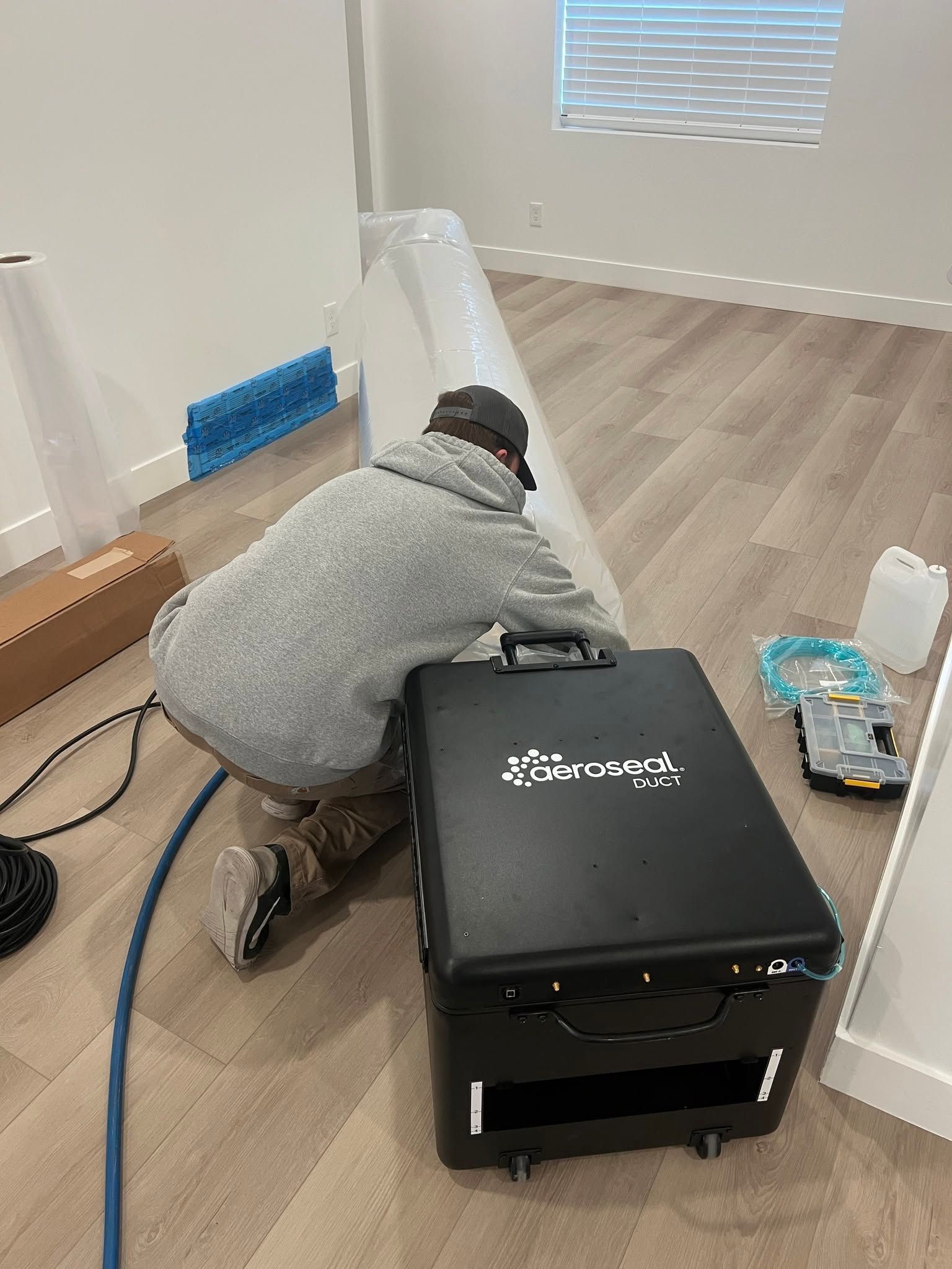 Person kneeling by a black machine on a hardwood floor in a bright room, working with cables and tools