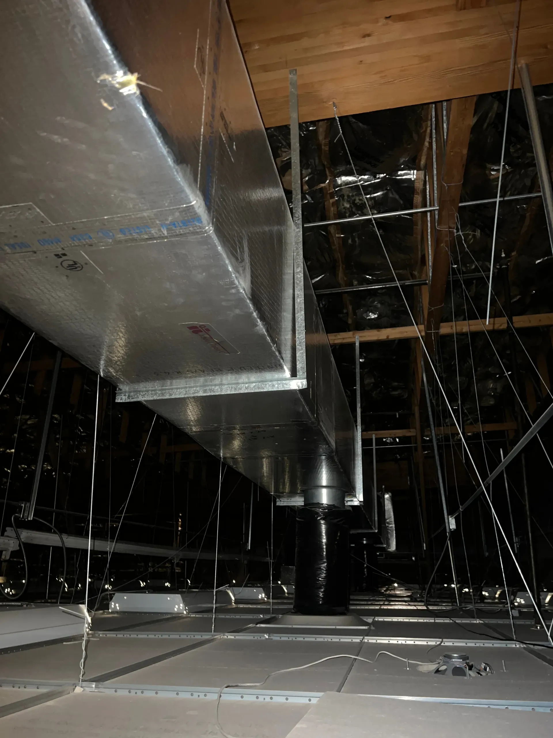 Exposed HVAC ductwork in a dark attic, with pipes, rafters, and hanging wires visible.