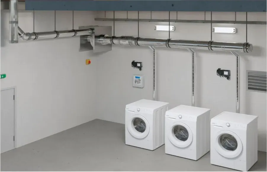 Empty laundry room with three white washers, gray walls, and exposed pipes along the ceiling
