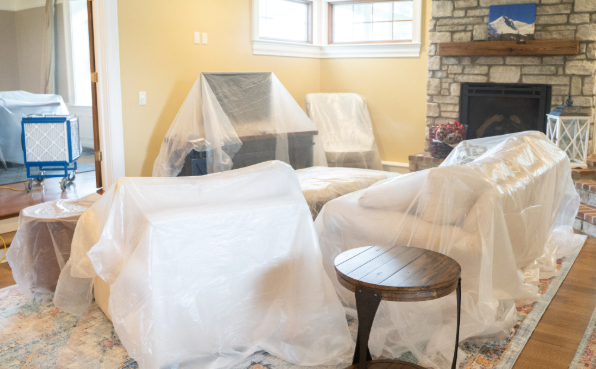 Living room with furniture covered in plastic during renovation, near a fireplace and window.